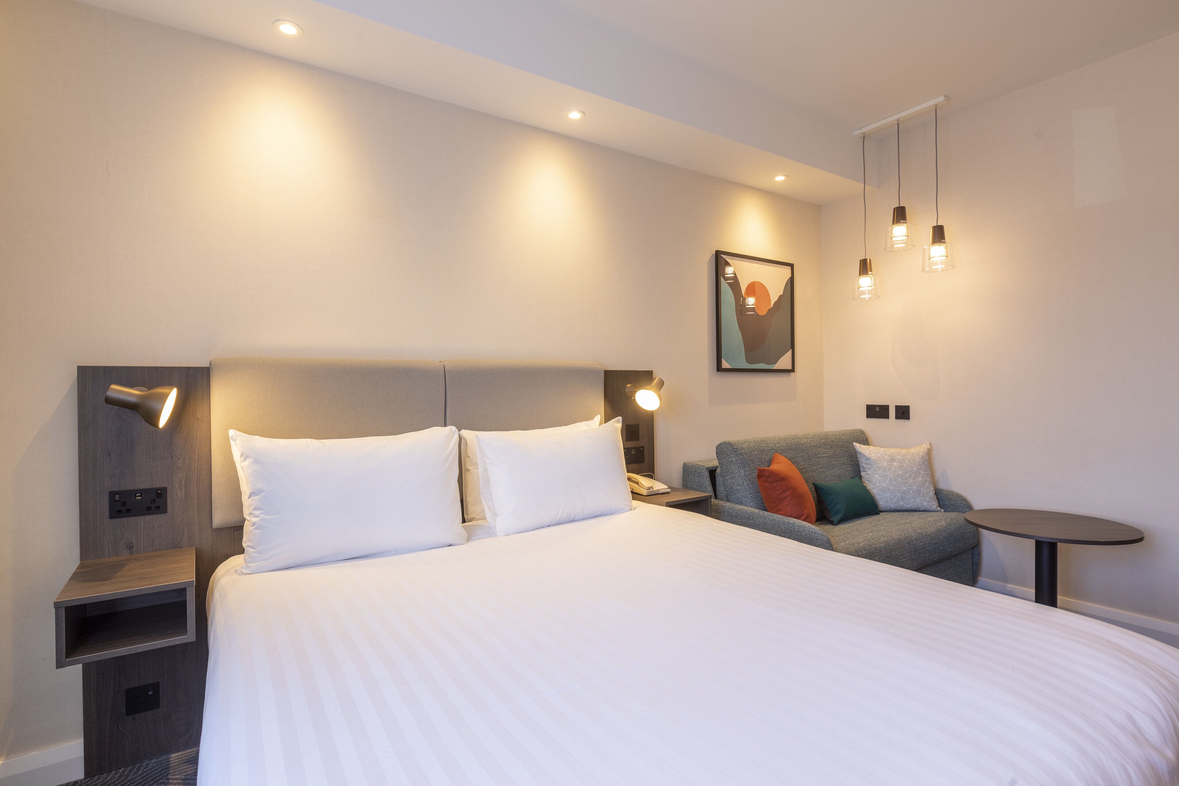 Une chambre d’hôtel moderne dotée d’un grand lit, d’un coin salon avec un canapé et un fauteuil, d’une petite table et d’un luminaire fixé au mur.