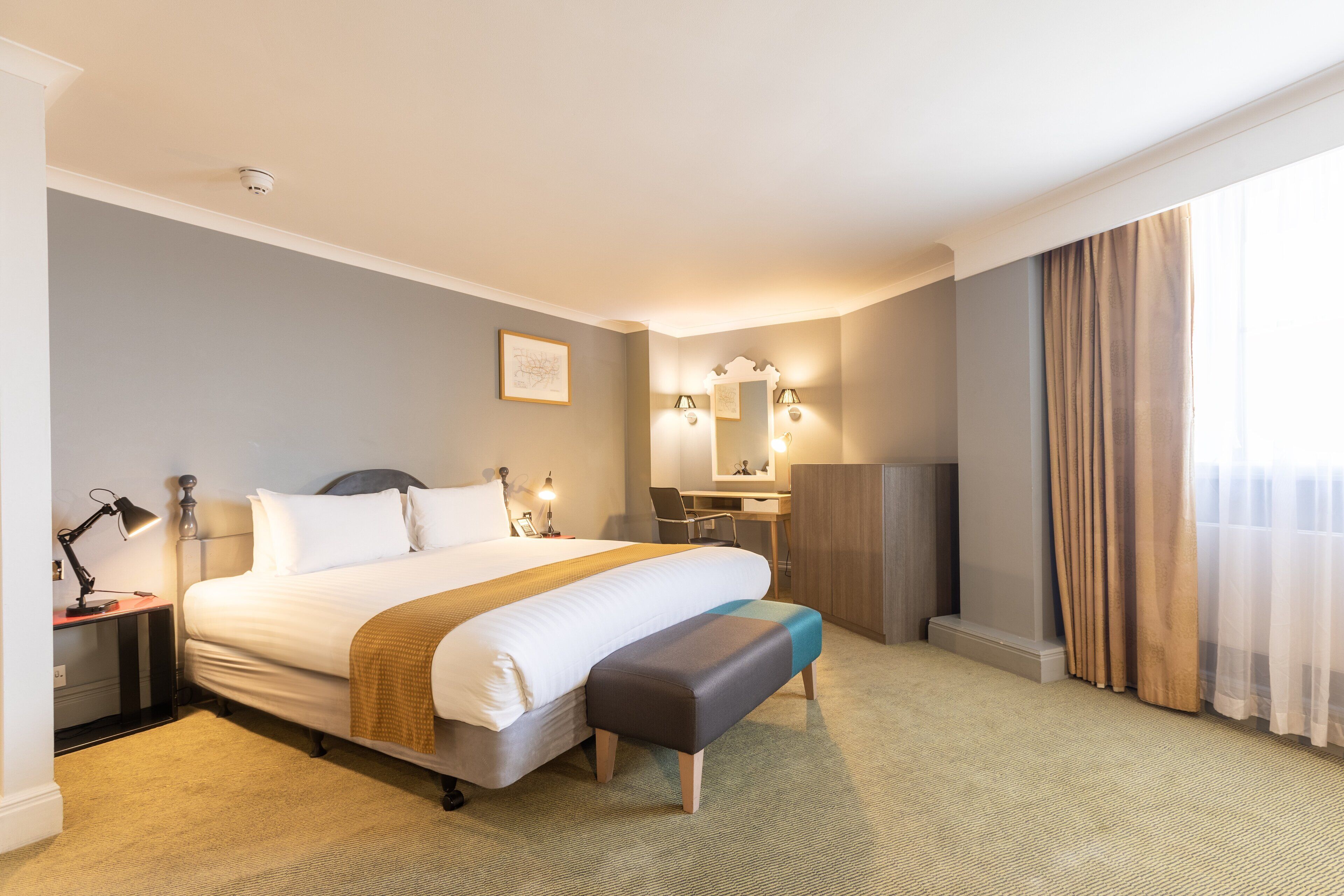 A hotel room with a large bed, bedside table, lamp, bench, desk, chair, and a large window with curtains.