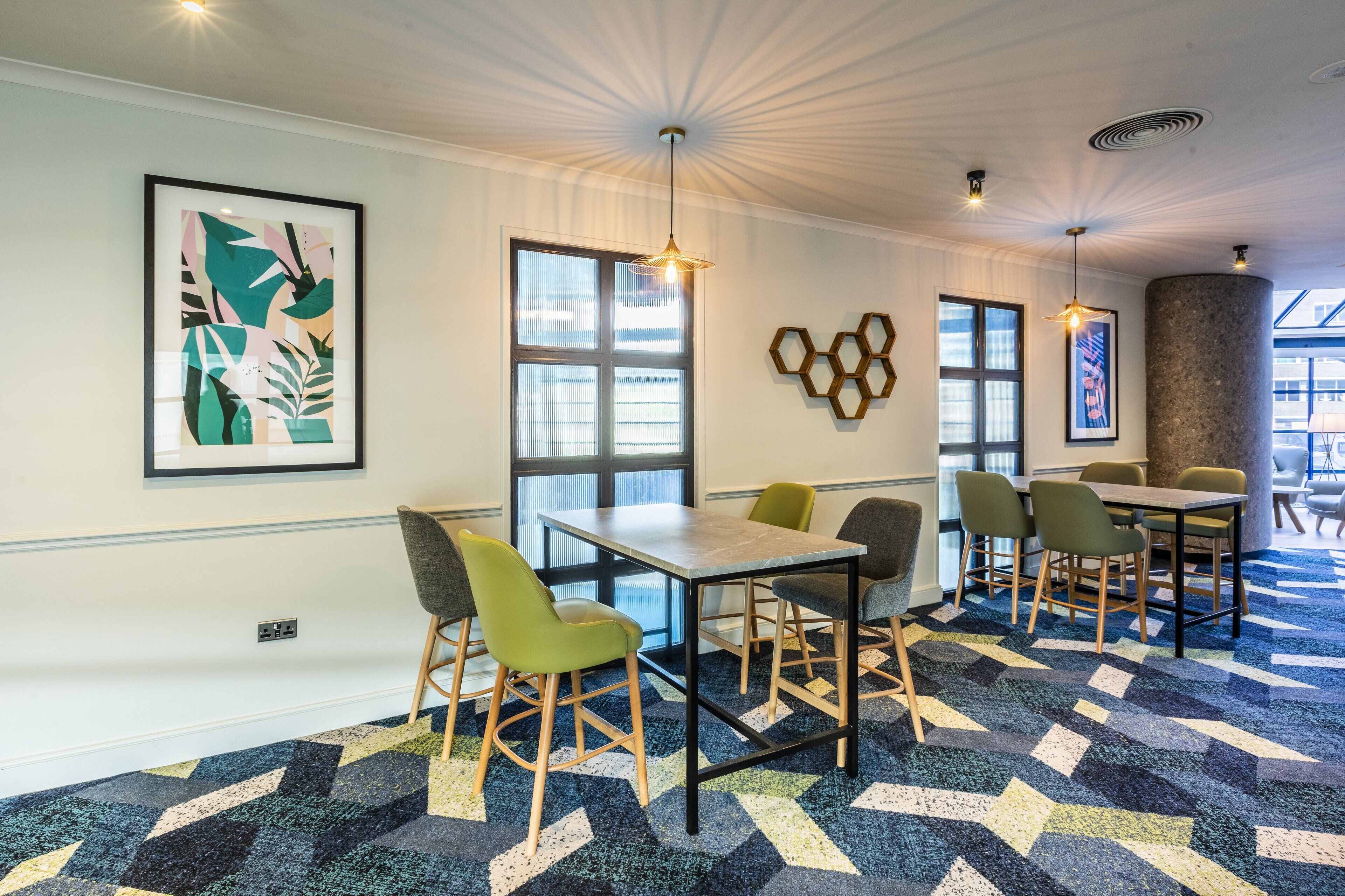 A modern conference room with a geometric carpet, green chairs, and a large window with blinds.