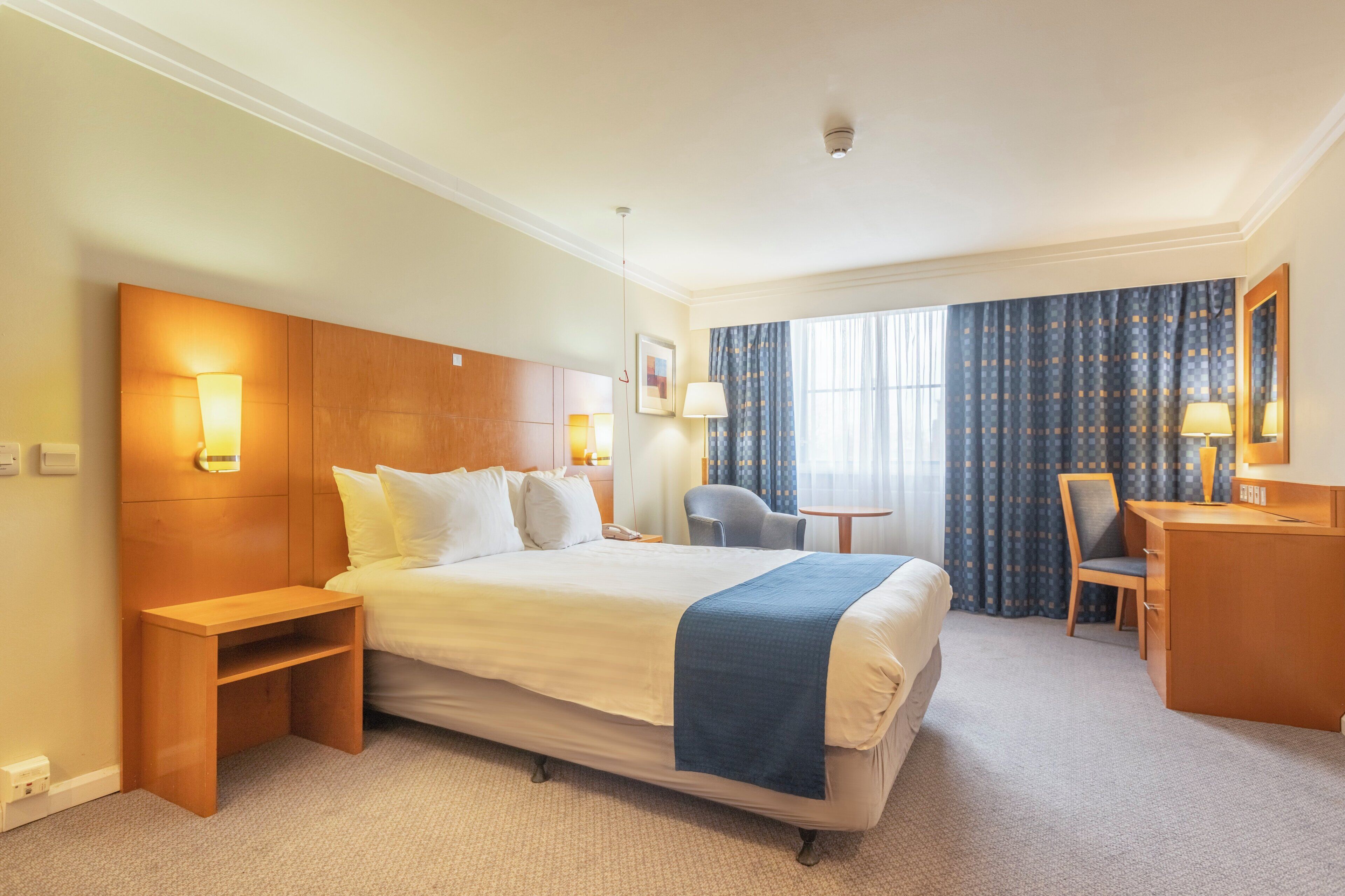 A hotel room with a large bed, bedside tables, a desk, a chair, and a window with curtains.