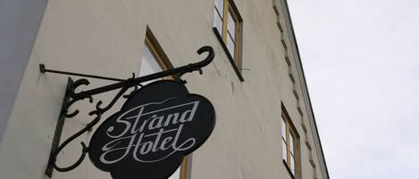 Een hotelsbord met de naam 'Grand Hotel' in een klassiek lettertype, bevestigd aan een gebouw met een witte gevel en meerdere ramen.