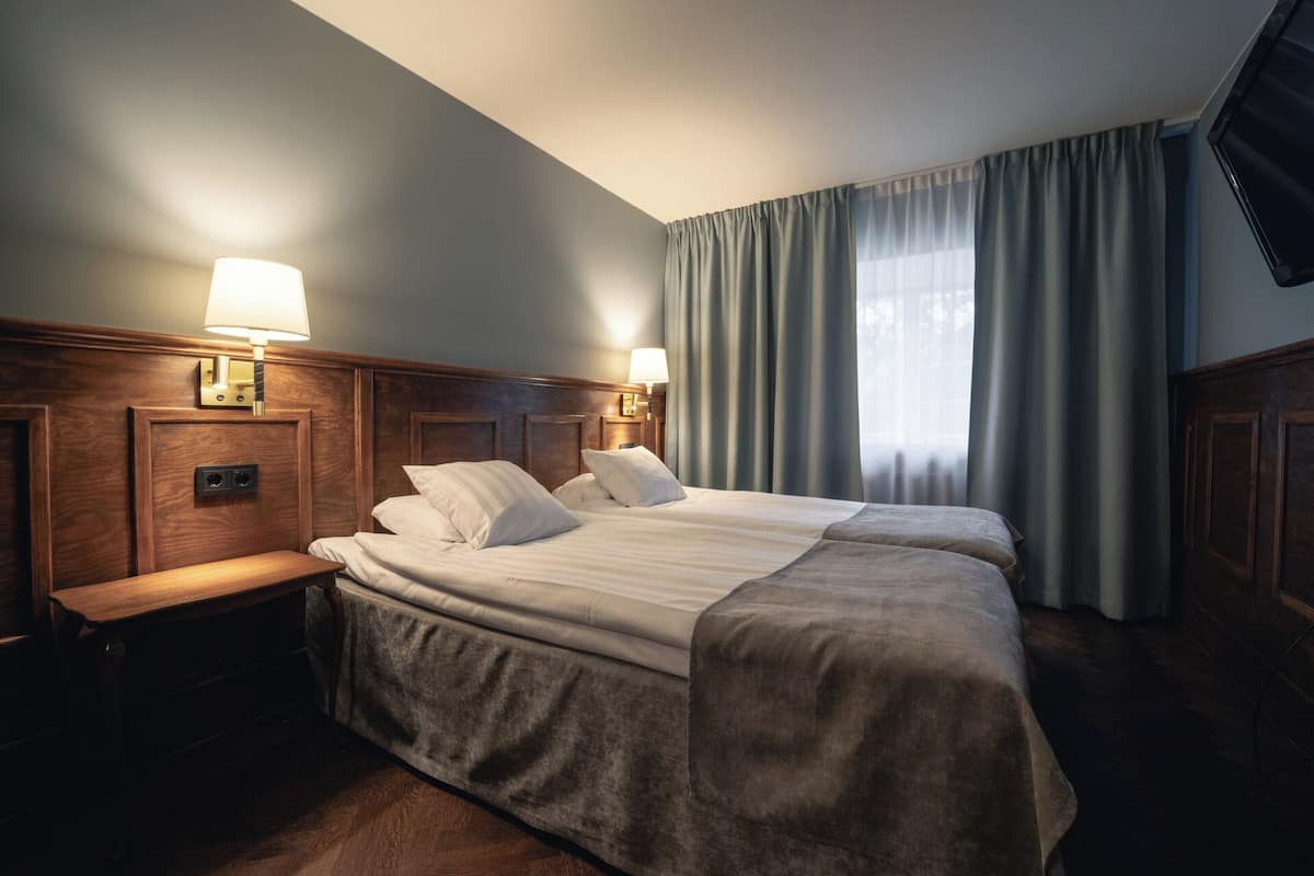 Une chambre d’hôtel avec un lit double, une tête de lit en bois, des lampes de chevet, un téléviseur à écran plat et une grande fenêtre avec des rideaux.