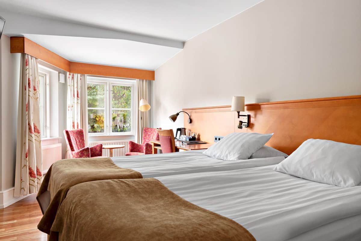 Une chambre d’hôtel avec deux lits, un coin salon comprenant une chaise et une table, et une fenêtre avec des rideaux.