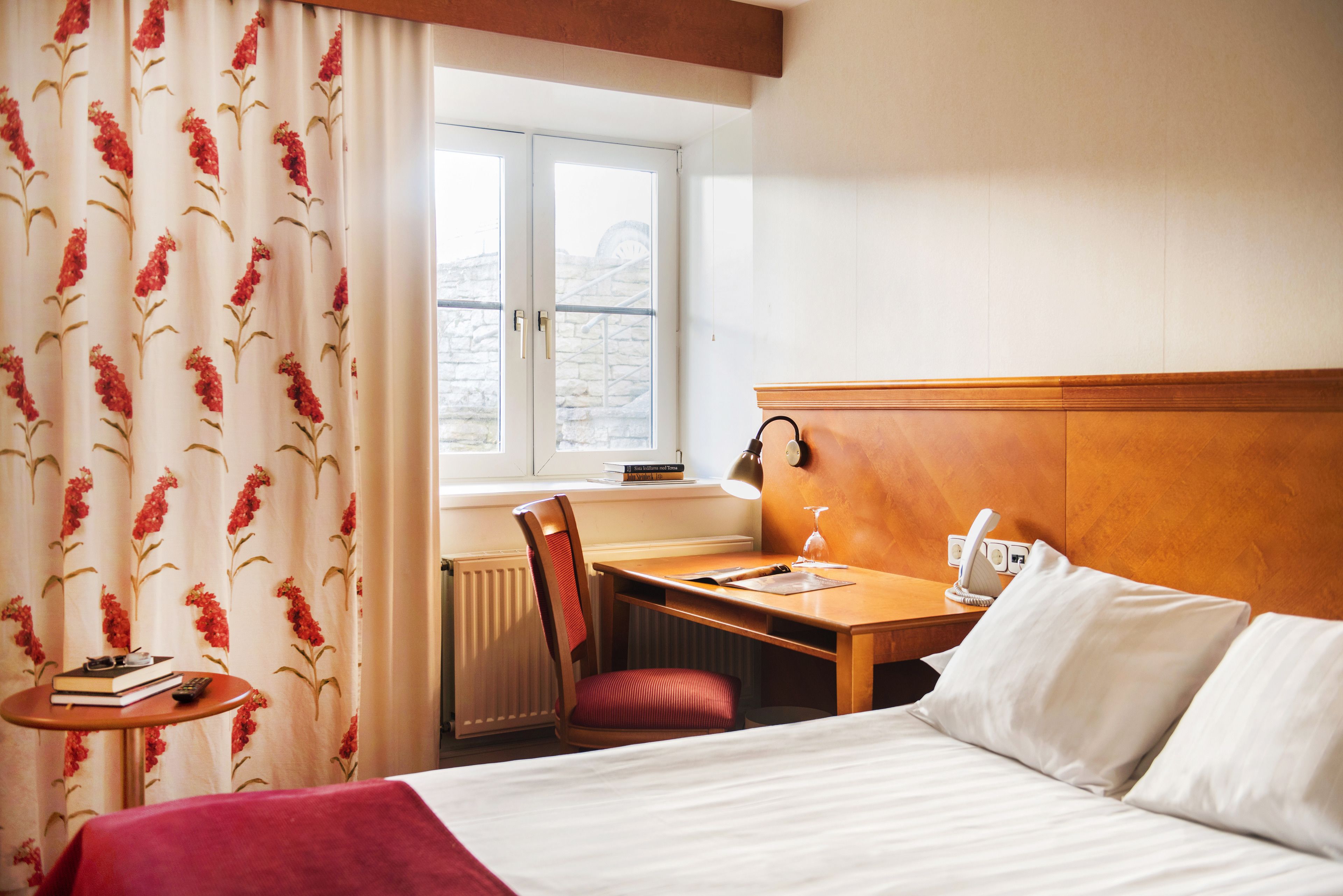 Une chambre d’hôtel comprenant un lit, un bureau, une chaise, une fenêtre avec des rideaux, un radiateur et une table de chevet avec une lampe et un téléphone.