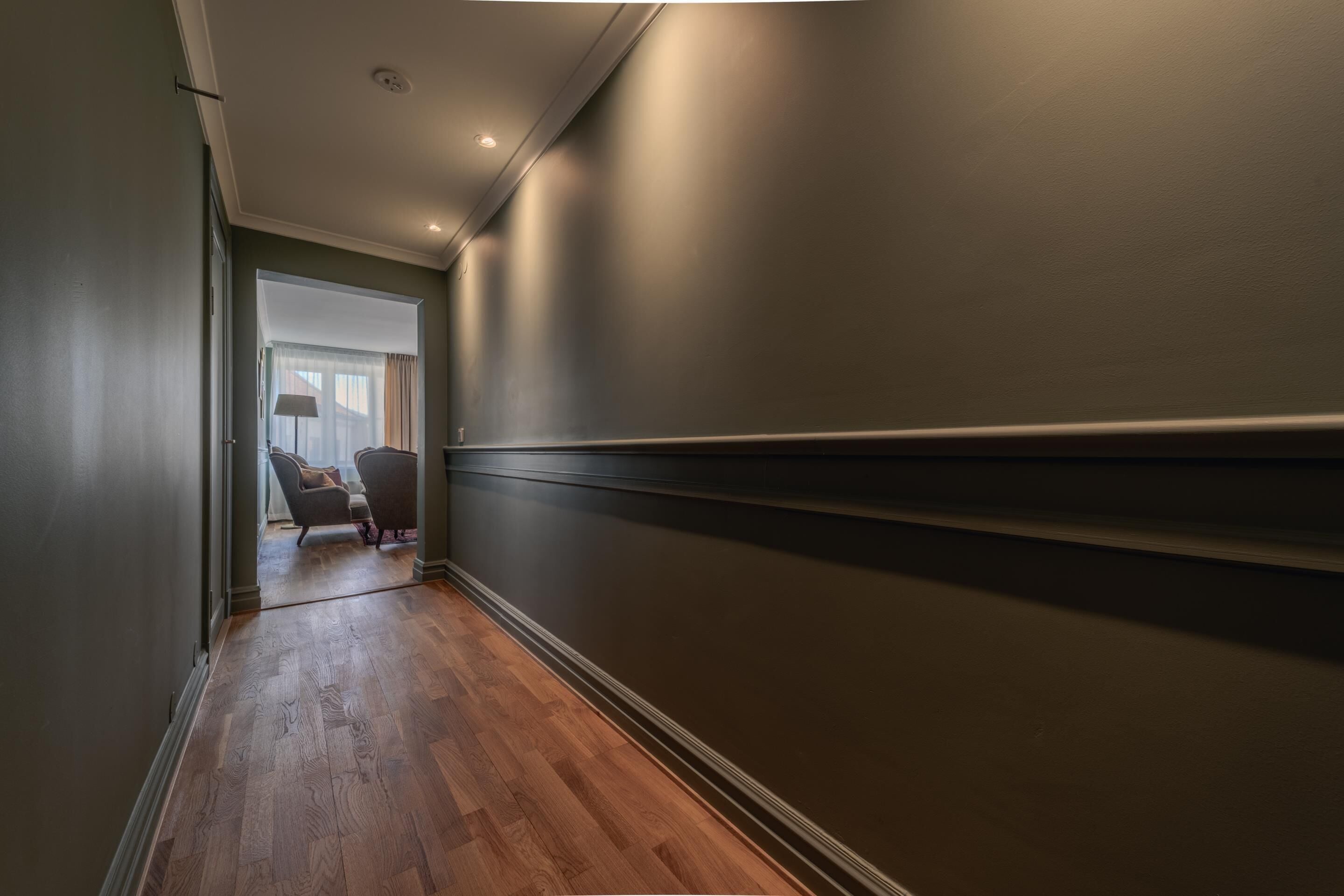 A hallway with wooden flooring, a chair, and a lamp near a window.