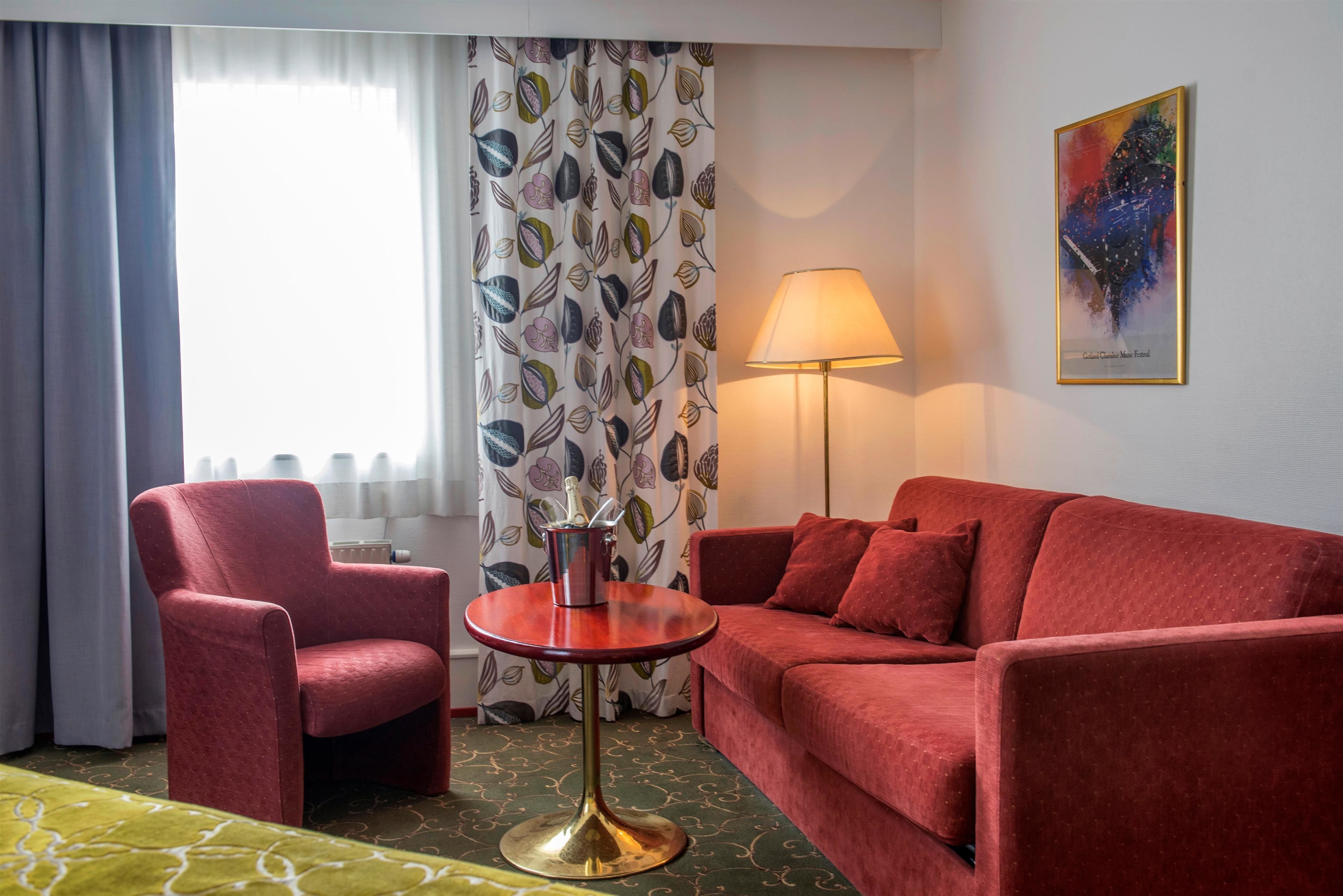 A hotel room with a red armchair, a red sofa, a small round table with a lamp, and a patterned curtain.