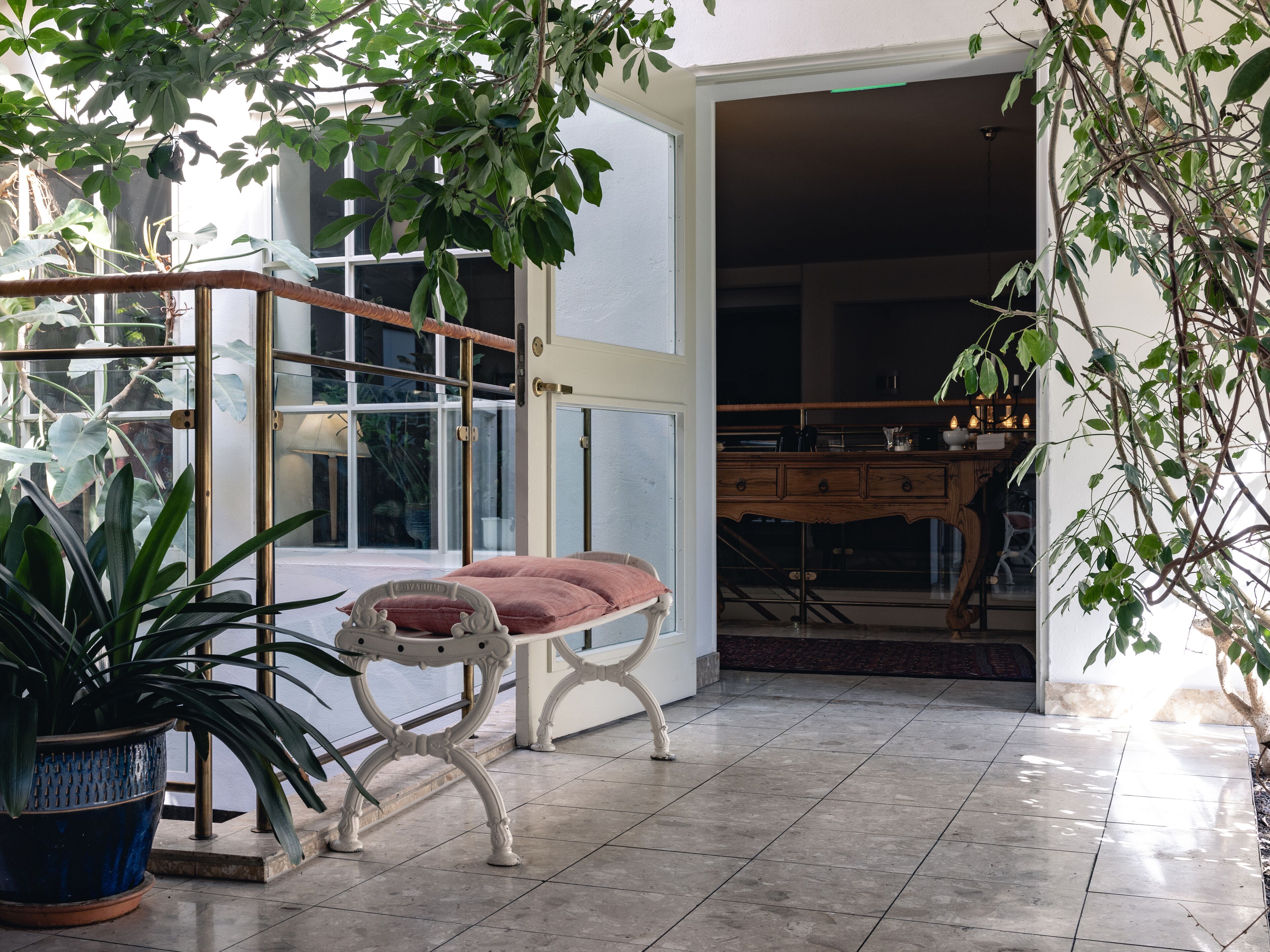 Una habitación con puerta de cristal que da a un balcón, un banco acolchado, una planta en maceta y un mueble de madera.