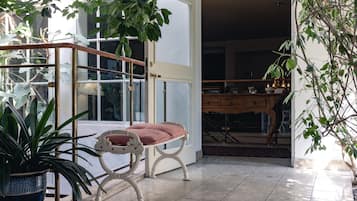 Una habitación con puerta de cristal que da a un balcón, un banco acolchado, una planta en maceta y un mueble de madera.