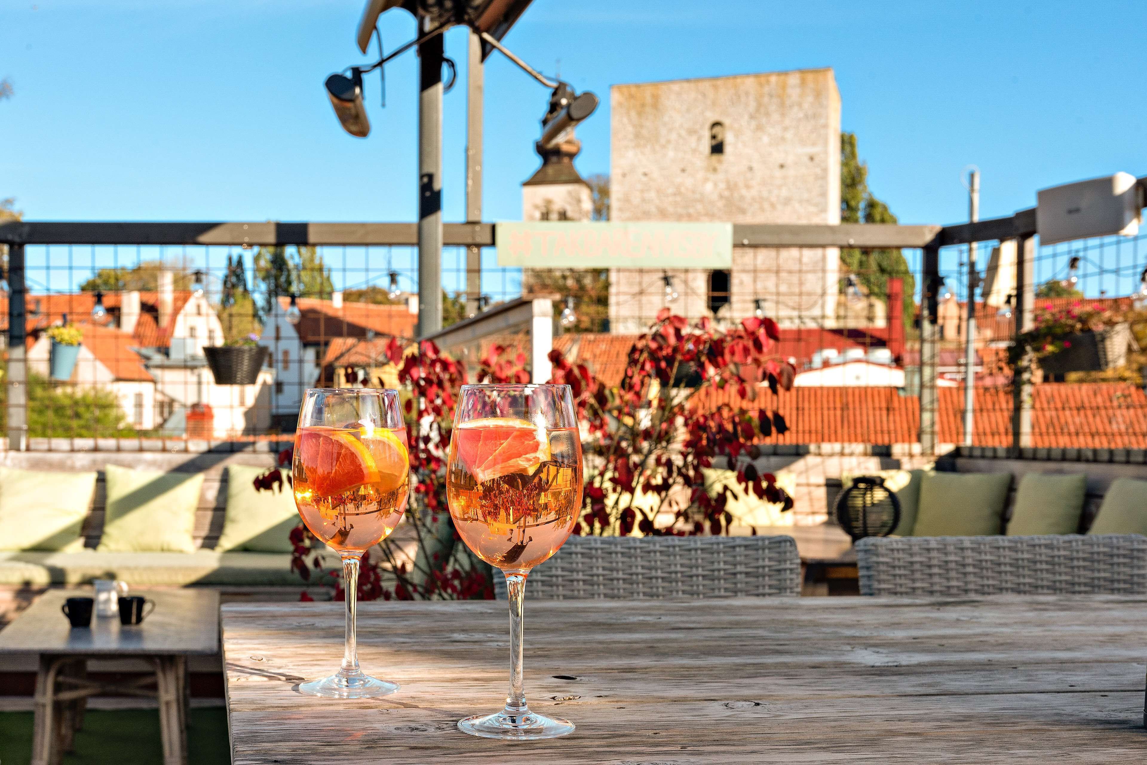 Deux verres de vin sur une table en bois, avec en arrière-plan une vue sur des bâtiments et une clôture.