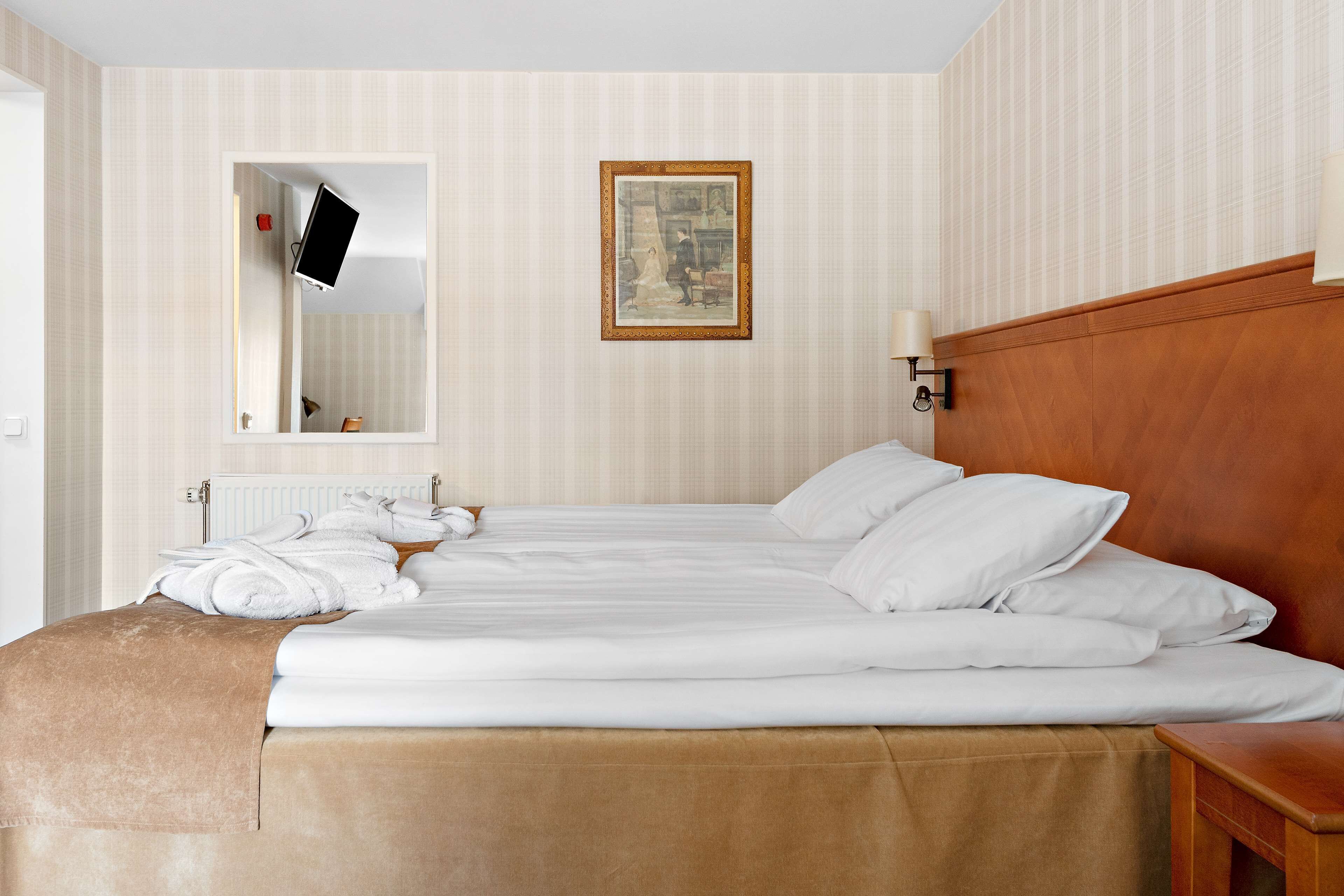 A neatly made bed with white linens, a wooden headboard, and a brown throw blanket. A flat-screen TV is mounted on the wall above the bed, and a framed picture hangs on the wall.