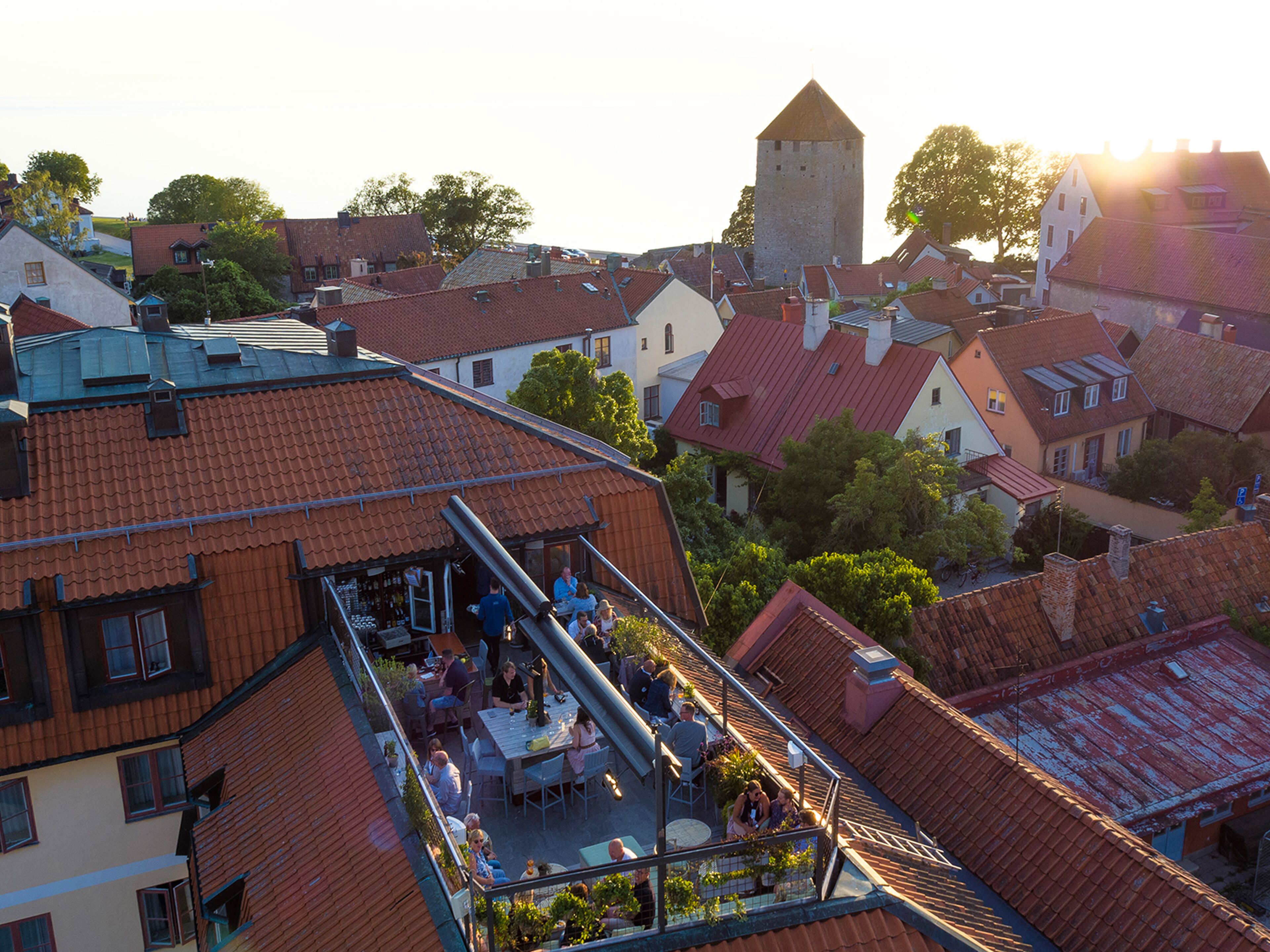 A rooftop terrace with tables and chairs, surrounded by buildings with red-tiled roofs and a prominent tower.