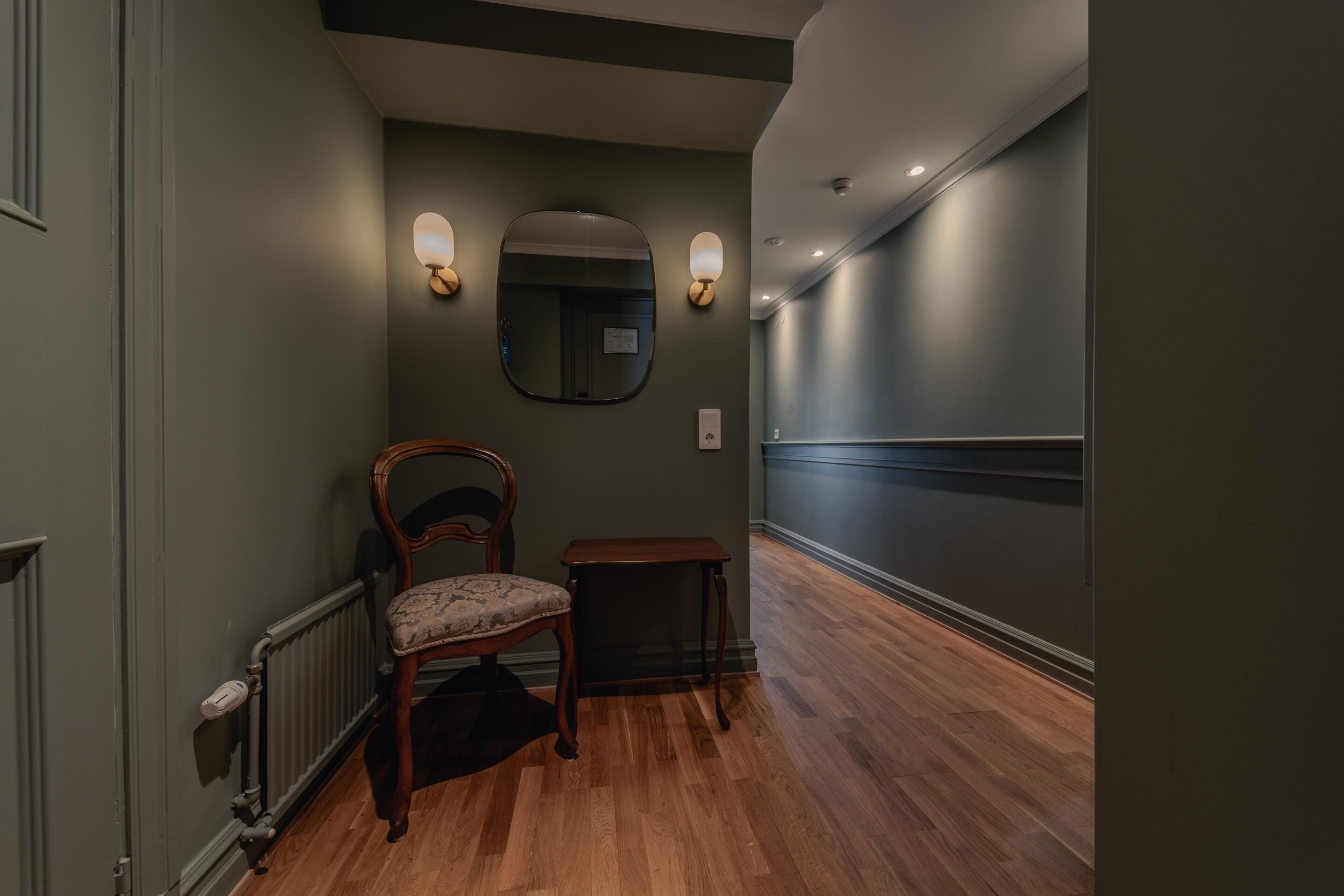 Un pasillo con suelo de madera, una silla, una mesita y apliques en la pared.