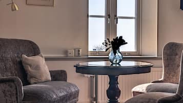 Ein Zimmer mit zwei Sesseln, einem kleinen Tisch mit einer Vase, einem Fenster mit Jalousien und einer Heizung.