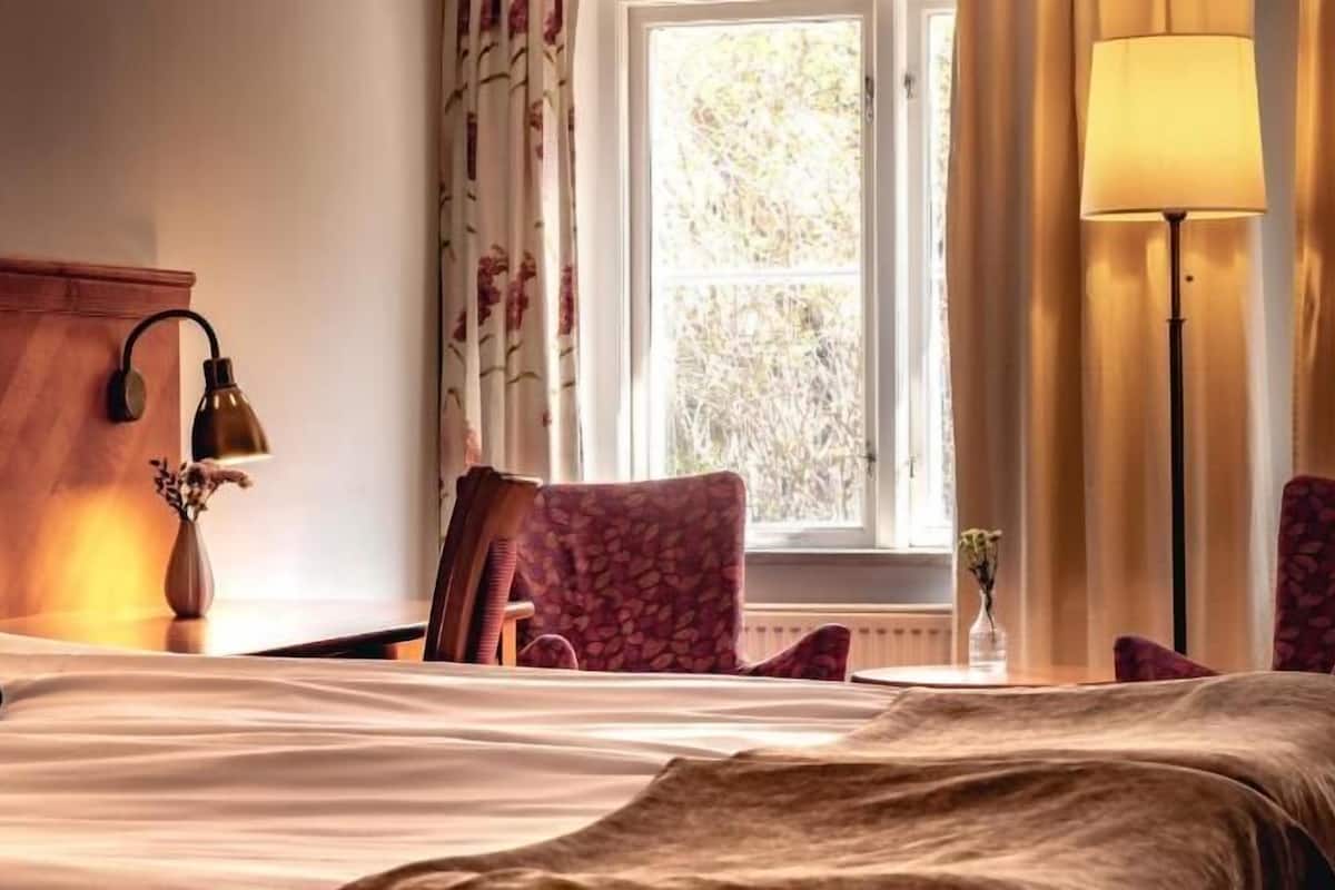 Une chambre d’hôtel avec un lit, une chaise, une lampe et une fenêtre avec des rideaux.