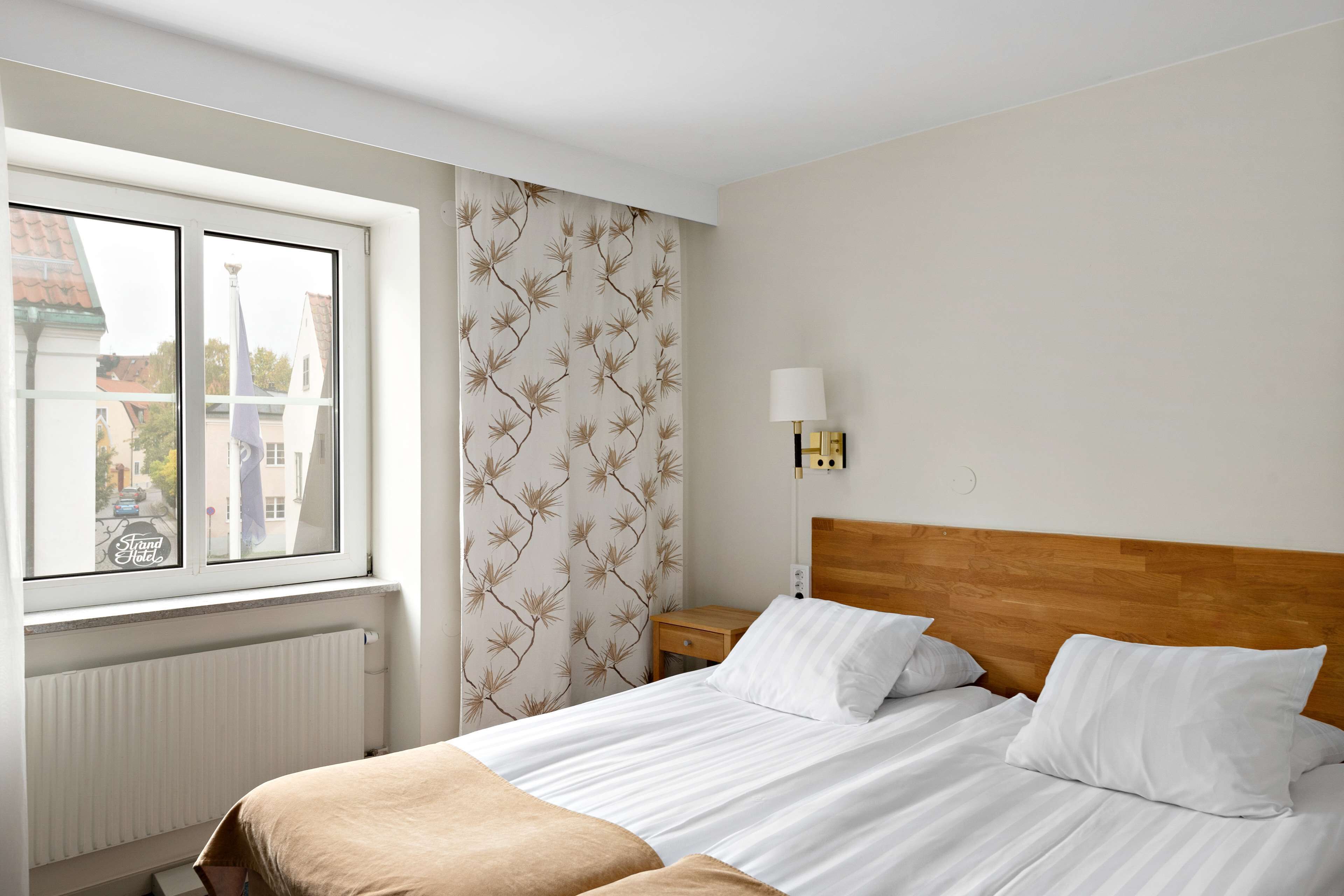 Quarto de hotel com cama, mesa de cabeceira, luminária e janela com vista para edifícios.