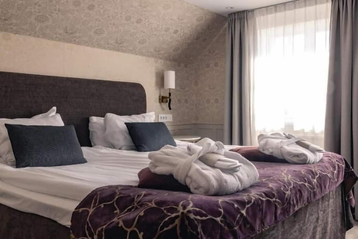Une chambre d’hôtel avec un grand lit, une tête de lit, une lampe de chevet et une vue sur la ville à travers des rideaux transparents.