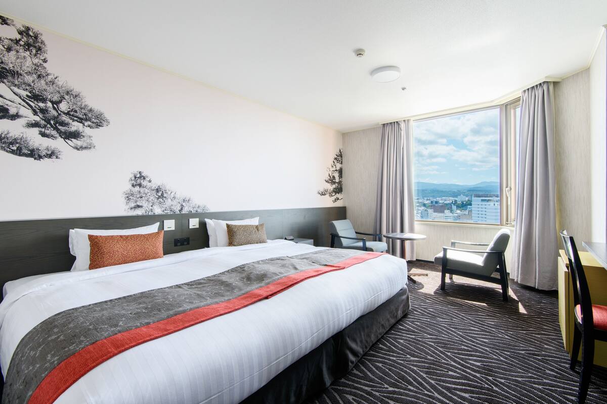Hotellihuone, jossa on suuri sänky, kaksi tuolia, pieni pöytä ja ikkunasta avautuva näkymä kaupunkiin.