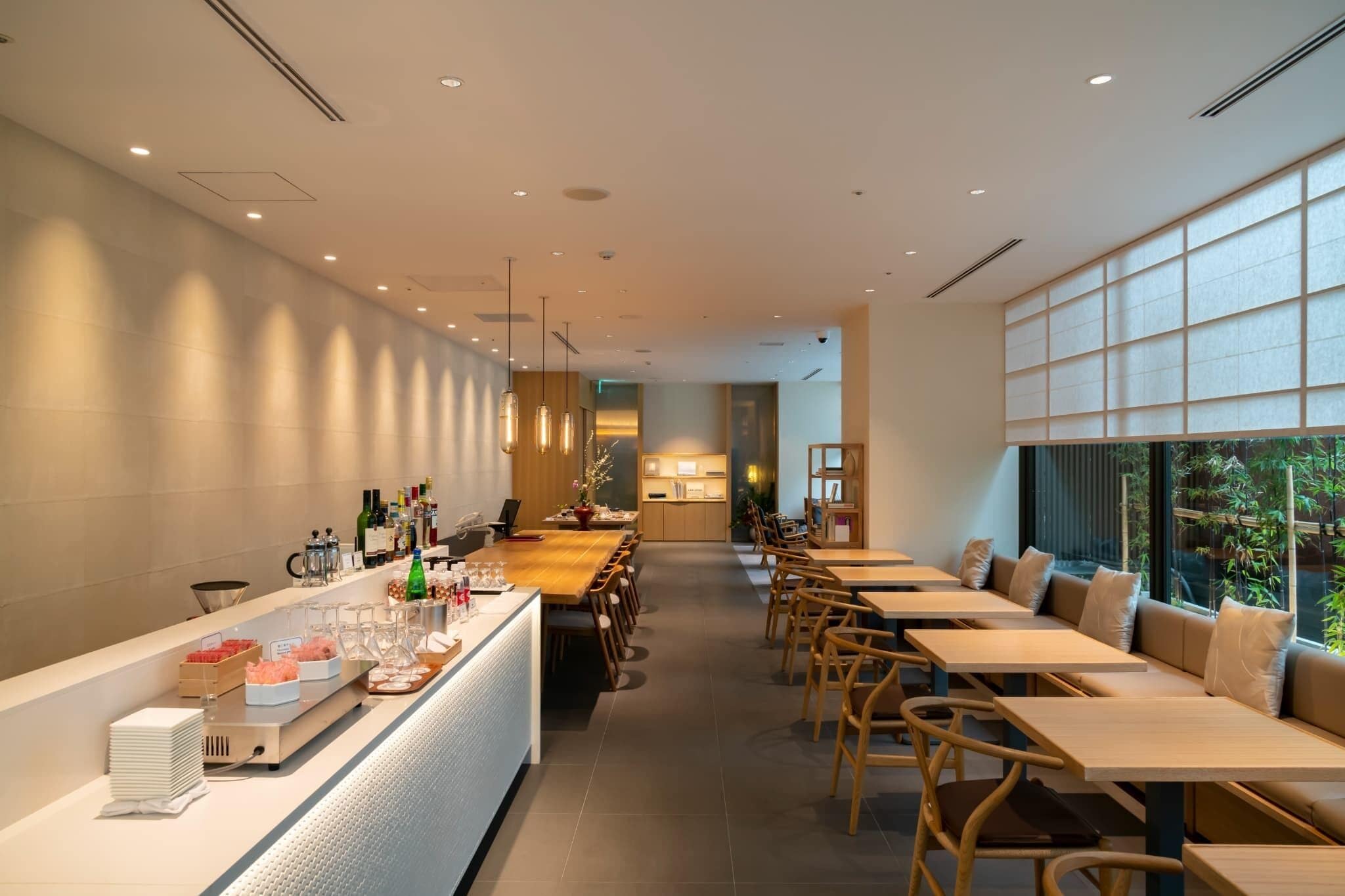 A modern restaurant with a long counter, wooden tables, and a view of greenery through large windows.