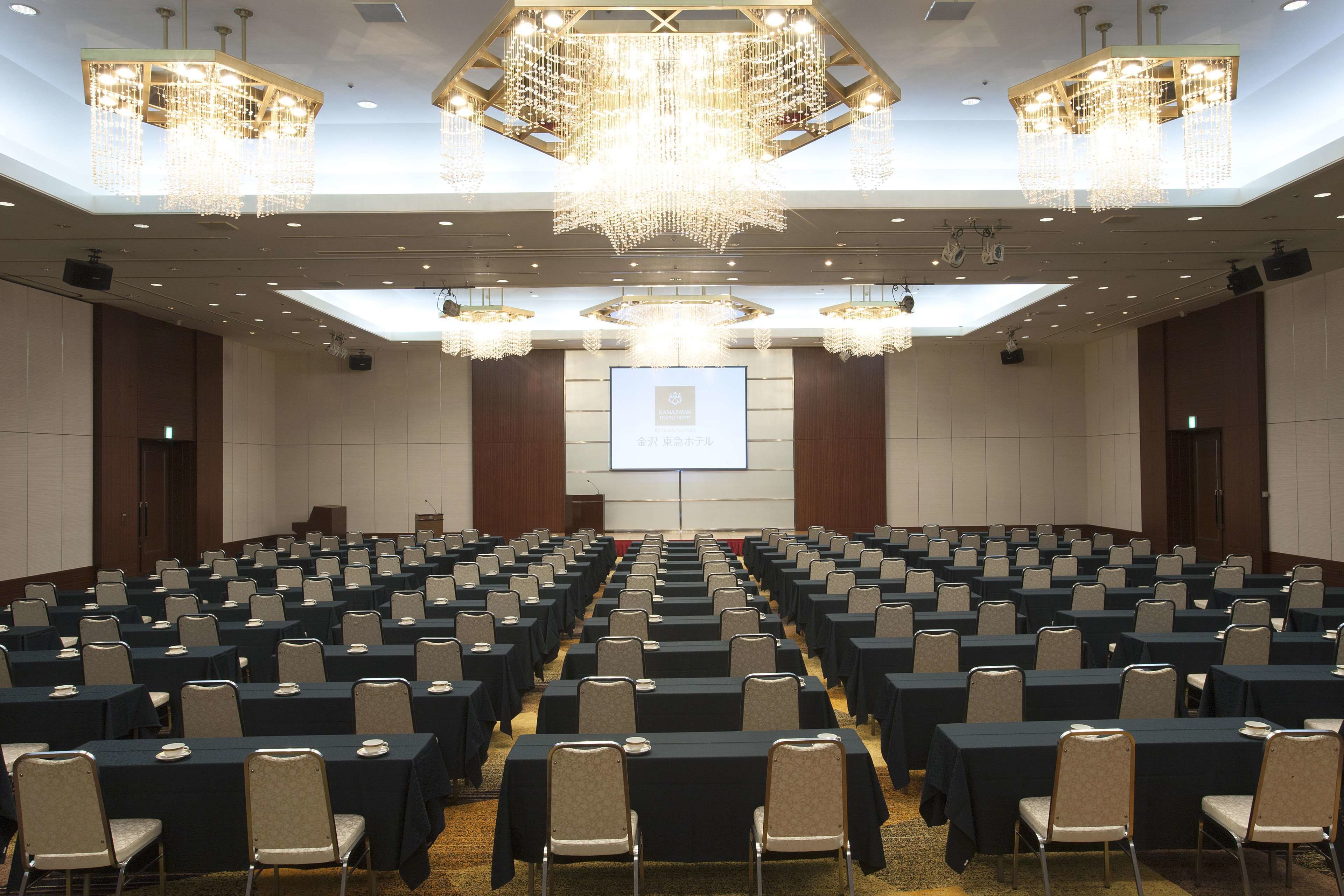 Un salón de conferencias con filas de mesas y sillas, una pantalla grande al frente y candelabros.