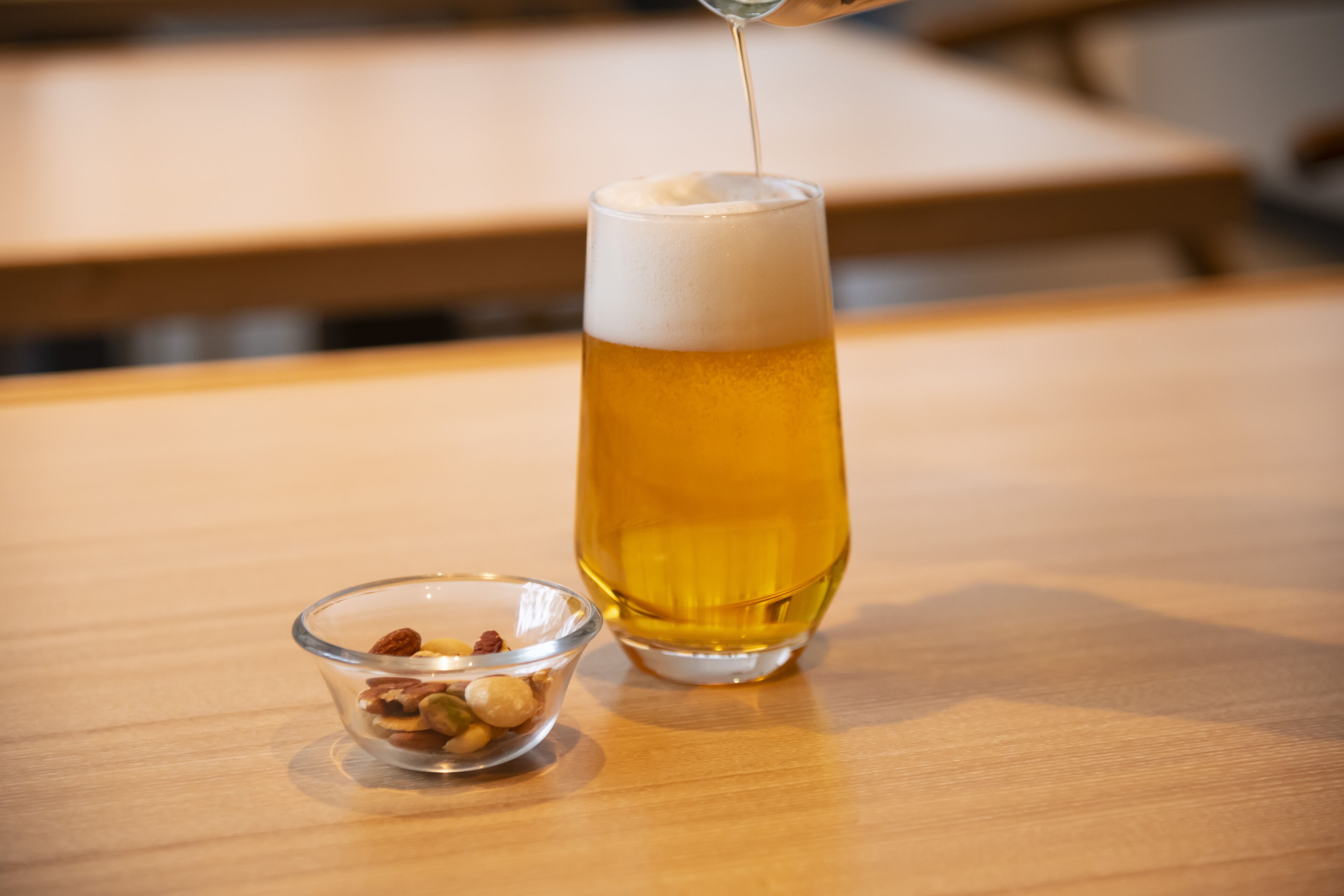 A glass of beer on a wooden table with a small bowl of nuts beside it.