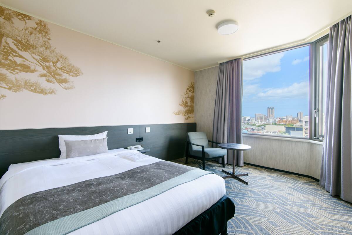 Hotellihuone, jossa on sänky, tuoli, pieni pöytä ja suuri ikkuna, josta avautuu näkymä kaupunkiin.