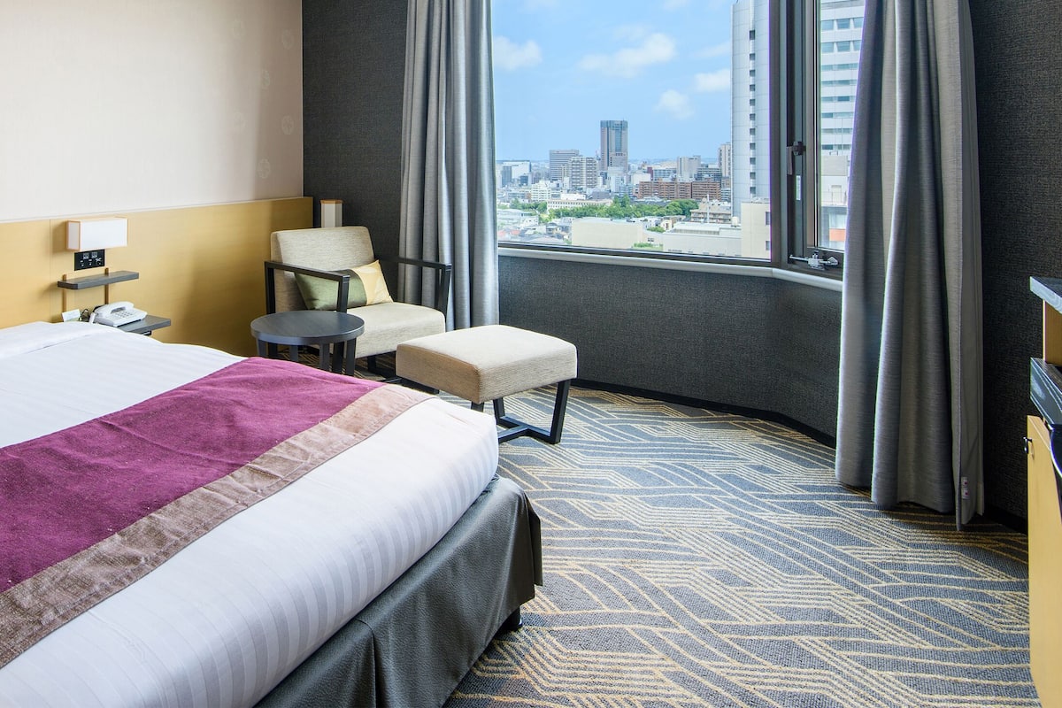 Hotellihuone, jossa on suuri sänky, tuoli, pieni pöytä ja näkymä kaupunkiin.