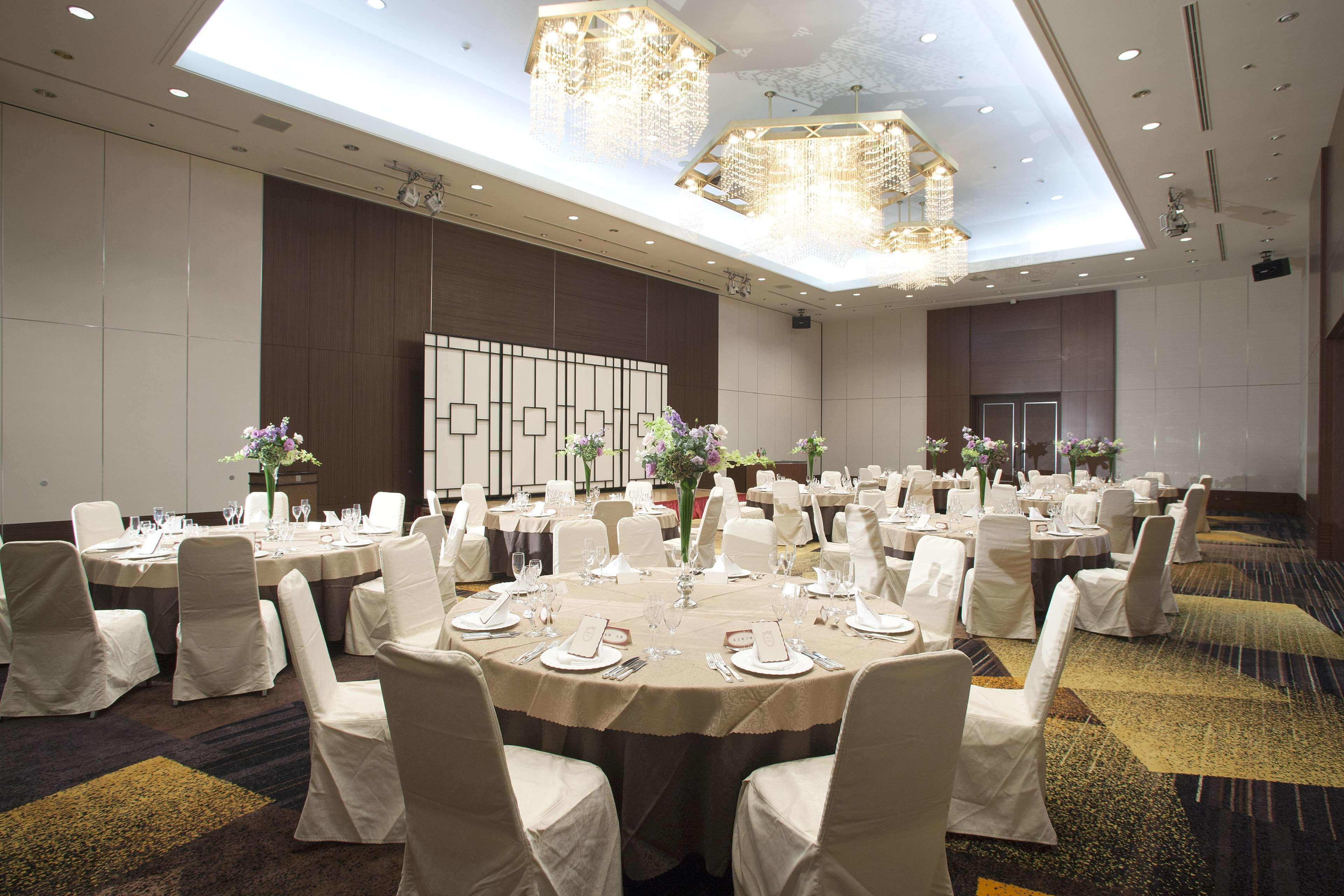 Uma sala de banquetes com mesas redondas dispostas para refeições, cadeiras brancas e lustres.