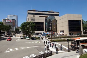 Una intersección urbana concurrida con edificios modernos, un paso de peatones y una parada de autobús.