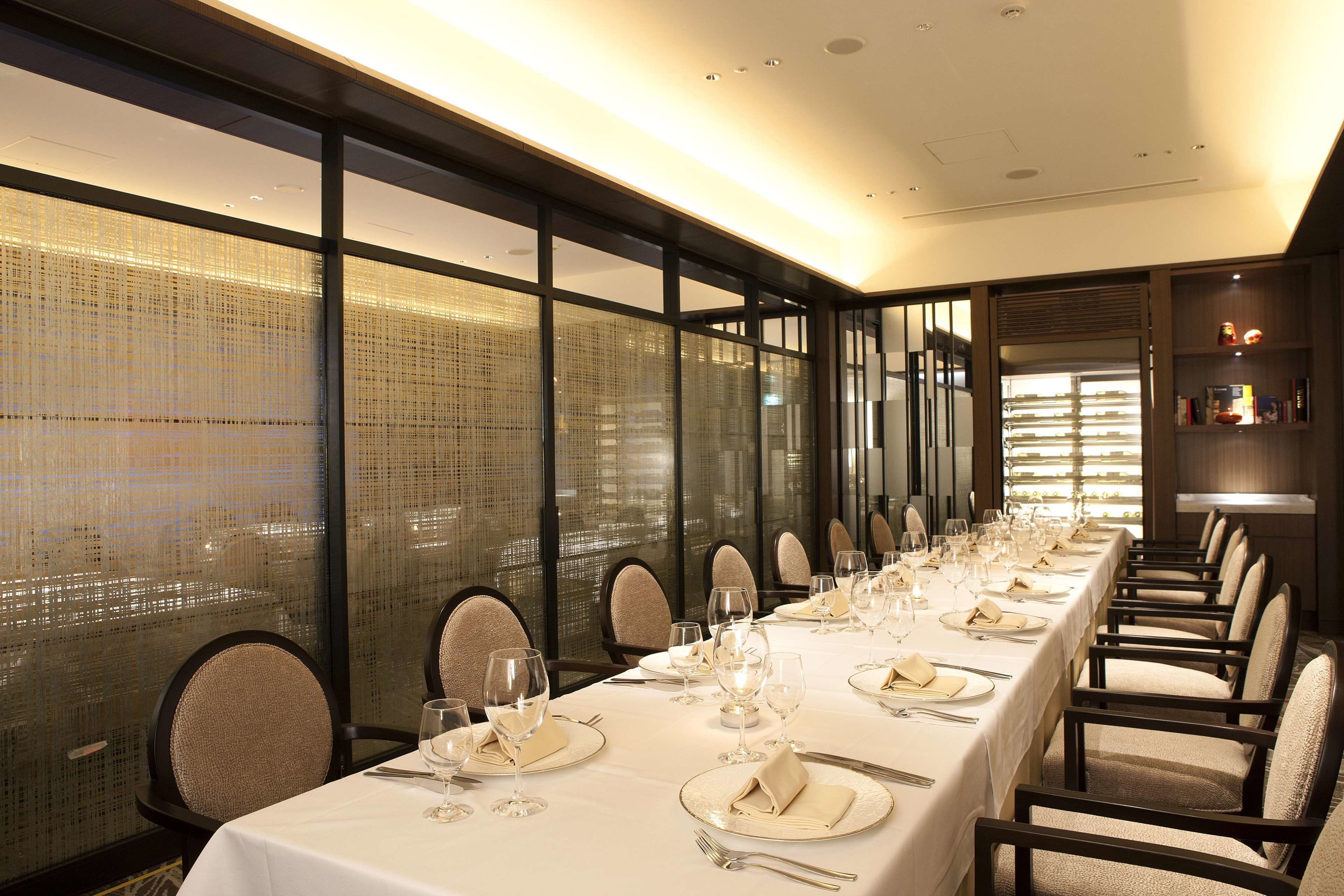 A long dining table set with white tablecloths, plates, and glasses, surrounded by chairs in a well-lit room with large windows.