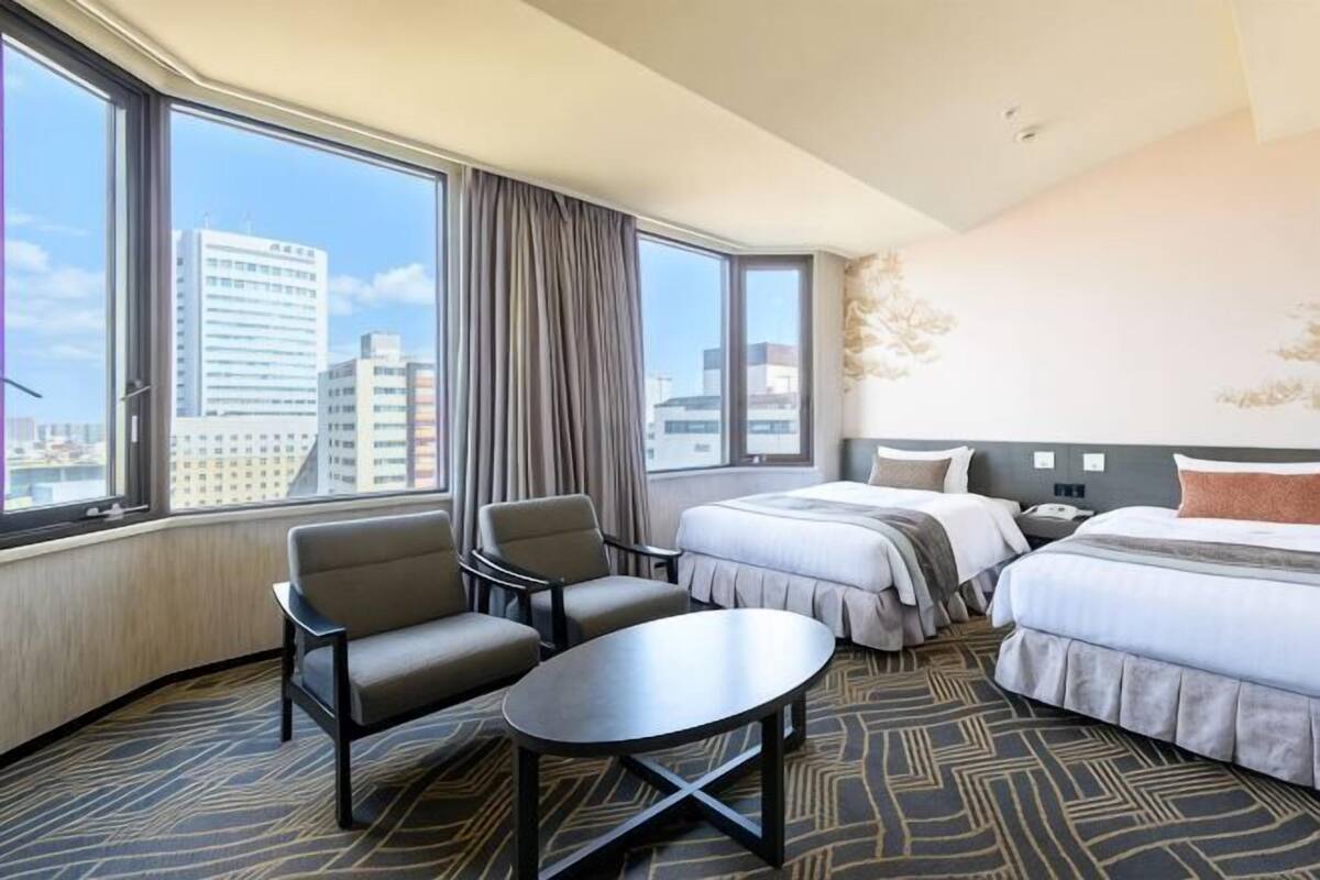 Hotellihuone, jossa on suuri ikkuna, kaksi sänkyä, työpöytä ja kaksi tuolia.