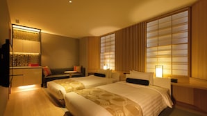 A hotel room with two beds, a sofa, a desk, and a large window with wooden blinds.