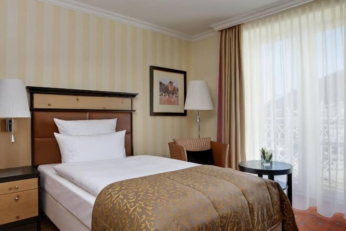 Une chambre d’hôtel avec un lit, des tables de chevet, des lampes, une petite table avec un vase et une fenêtre avec des rideaux.