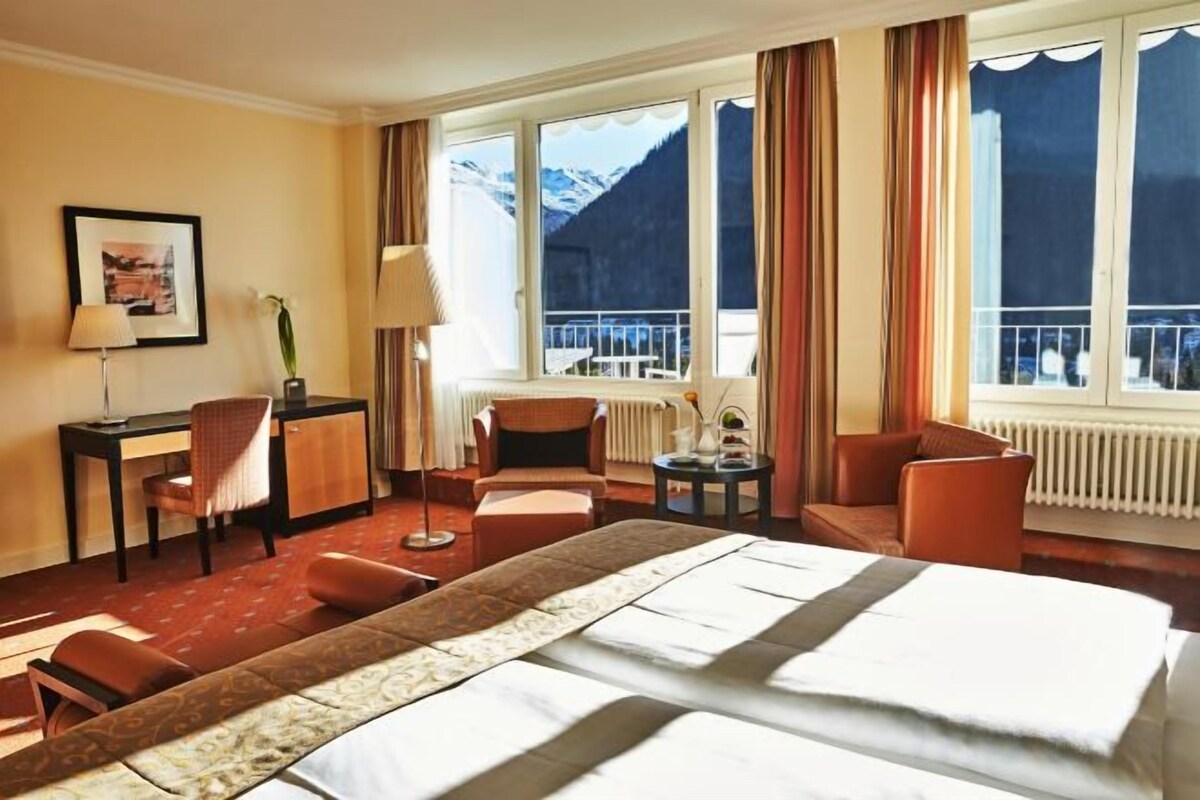 Habitación de hotel con una cama grande, un escritorio, dos sillas, una mesita y vistas a montañas a través de la ventana.