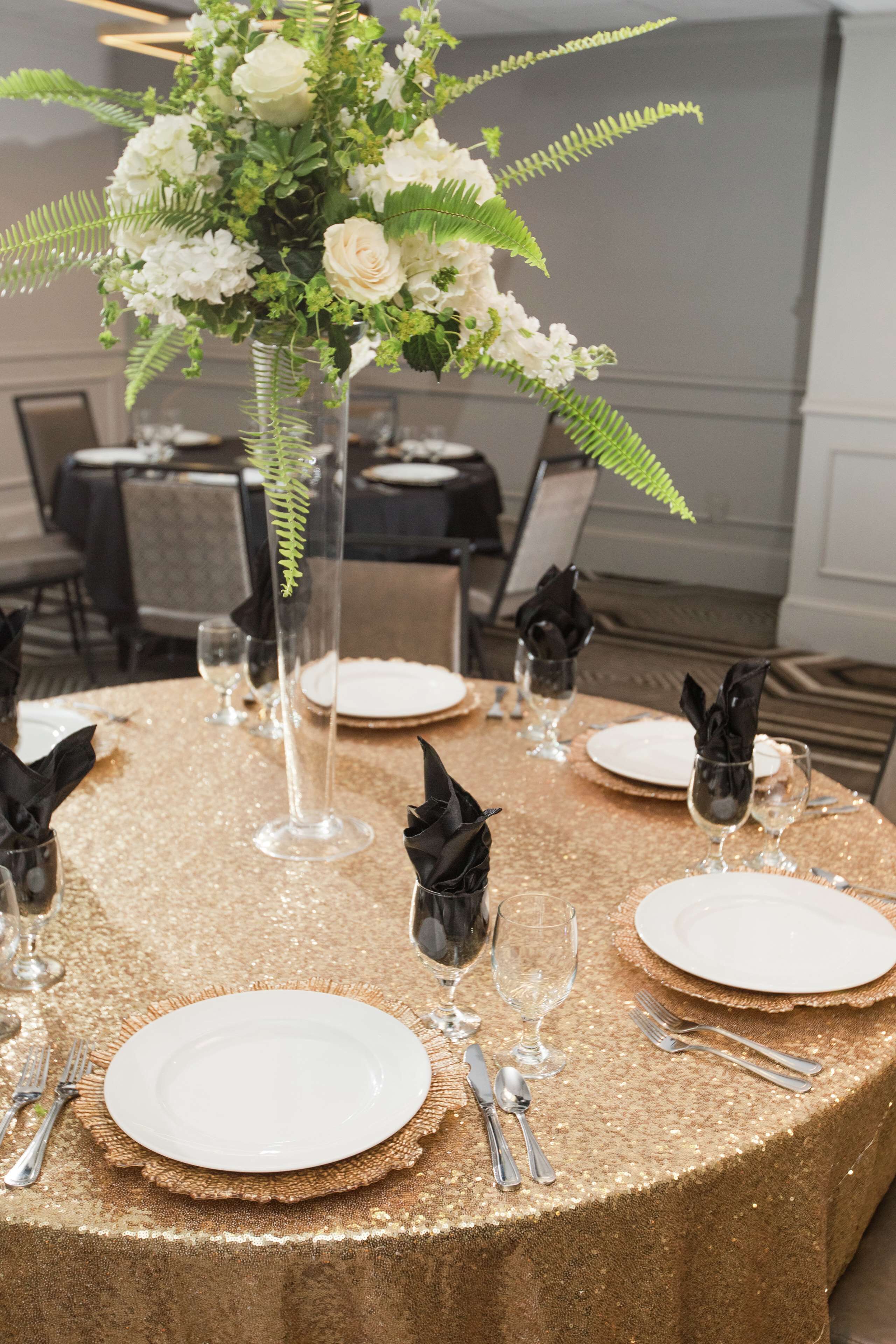 A round table with a gold sequin tablecloth, white plates, silverware, and black napkins. A tall vase with white flowers and green foliage is placed in the center.