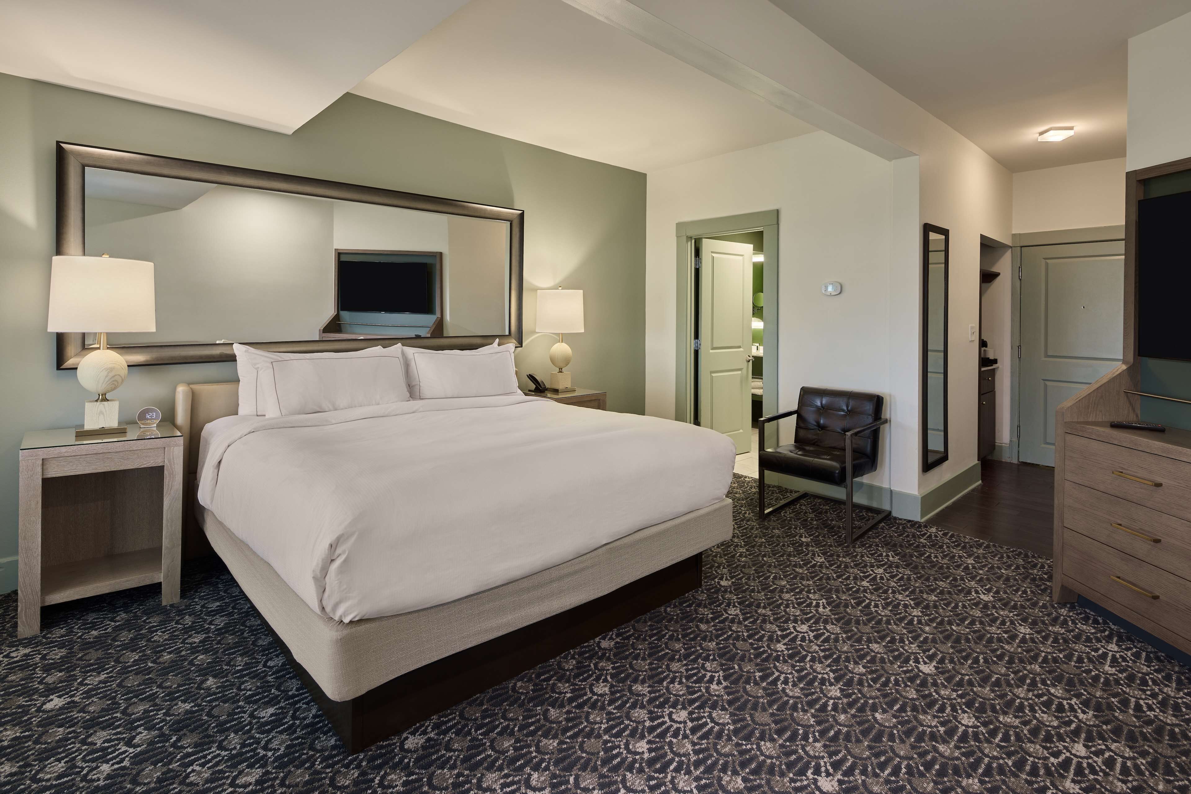 A hotel room with a large bed, two bedside tables with lamps, a chair, a dresser, and a large mirror above the bed.