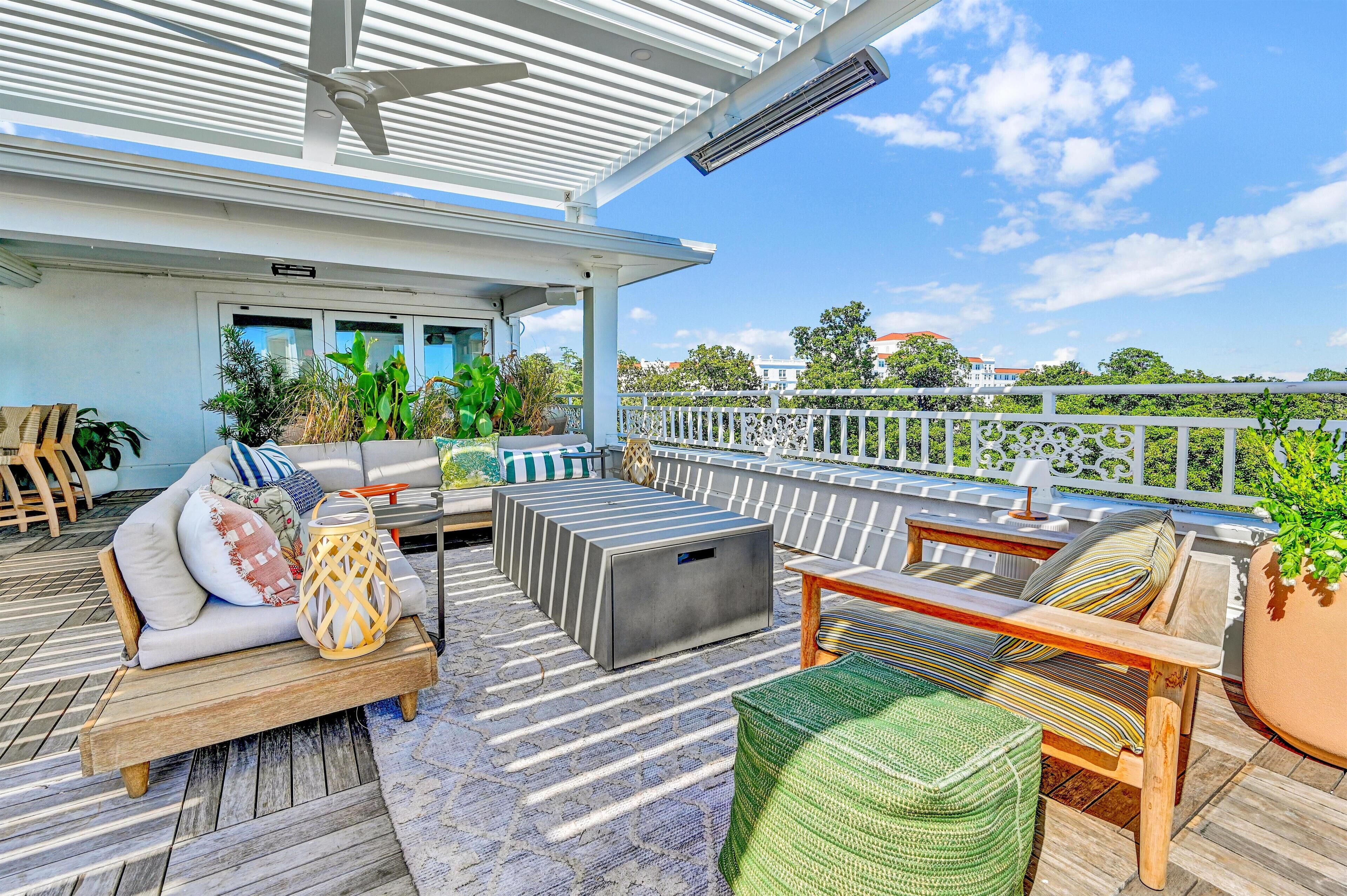 A covered outdoor seating area with a sofa, ottoman, and chairs, surrounded by potted plants and a view of the city.