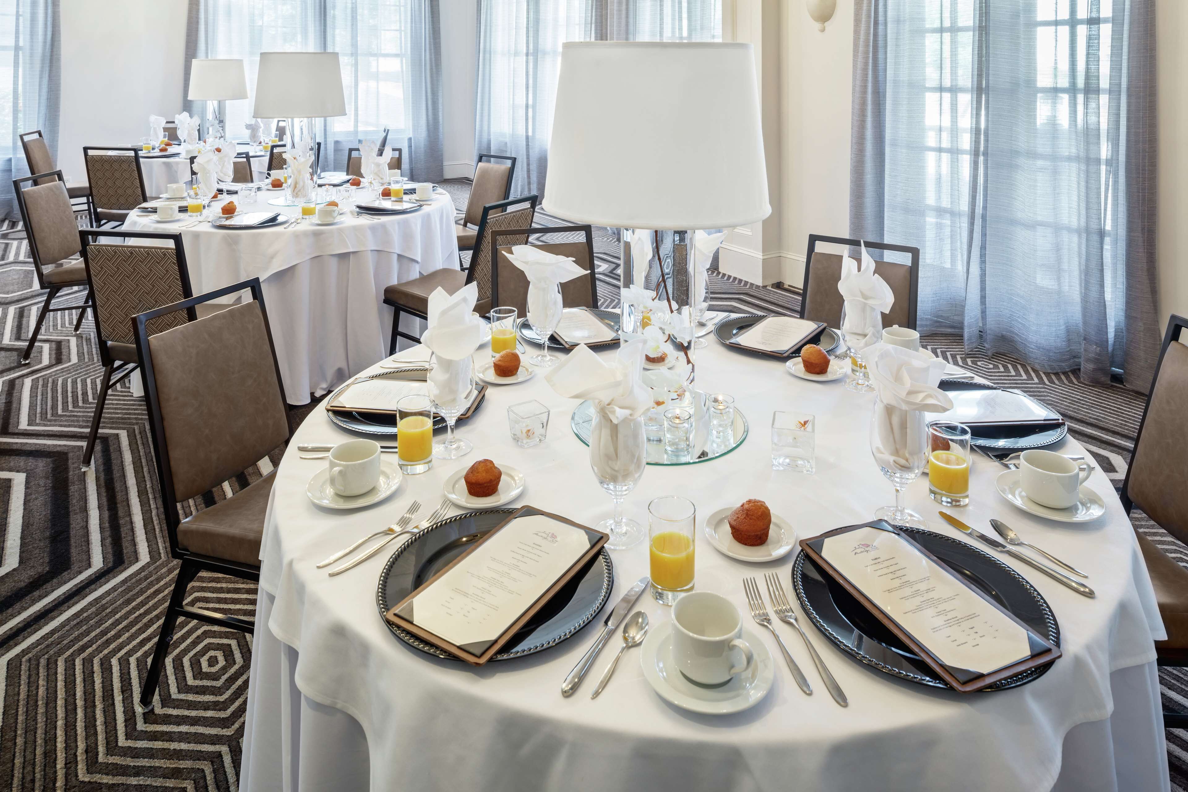 Une salle à manger d’hôtel avec des tables rondes dressées pour le petit-déjeuner, agrémentées de nappes blanches, de couverts en argent et de divers éléments pour le petit-déjeuner, comme des muffins et du jus d’orange.