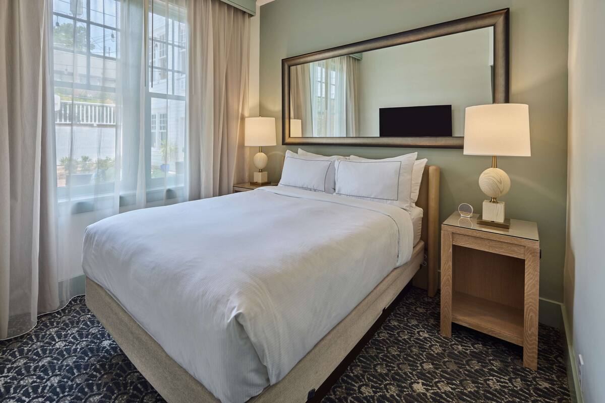 A hotel room with a large bed, two bedside tables with lamps, a mirror above the bed, and a window with curtains.