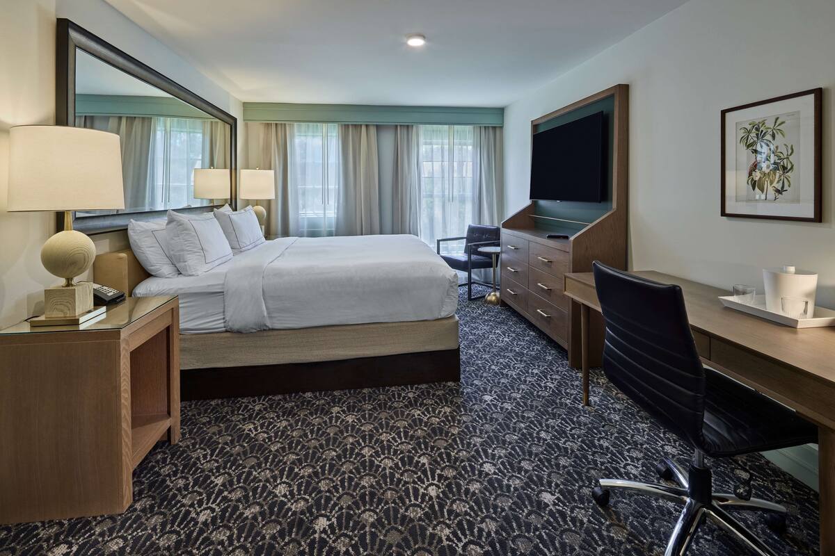 A hotel room with a large bed, a desk, a chair, a TV, and a mirror.