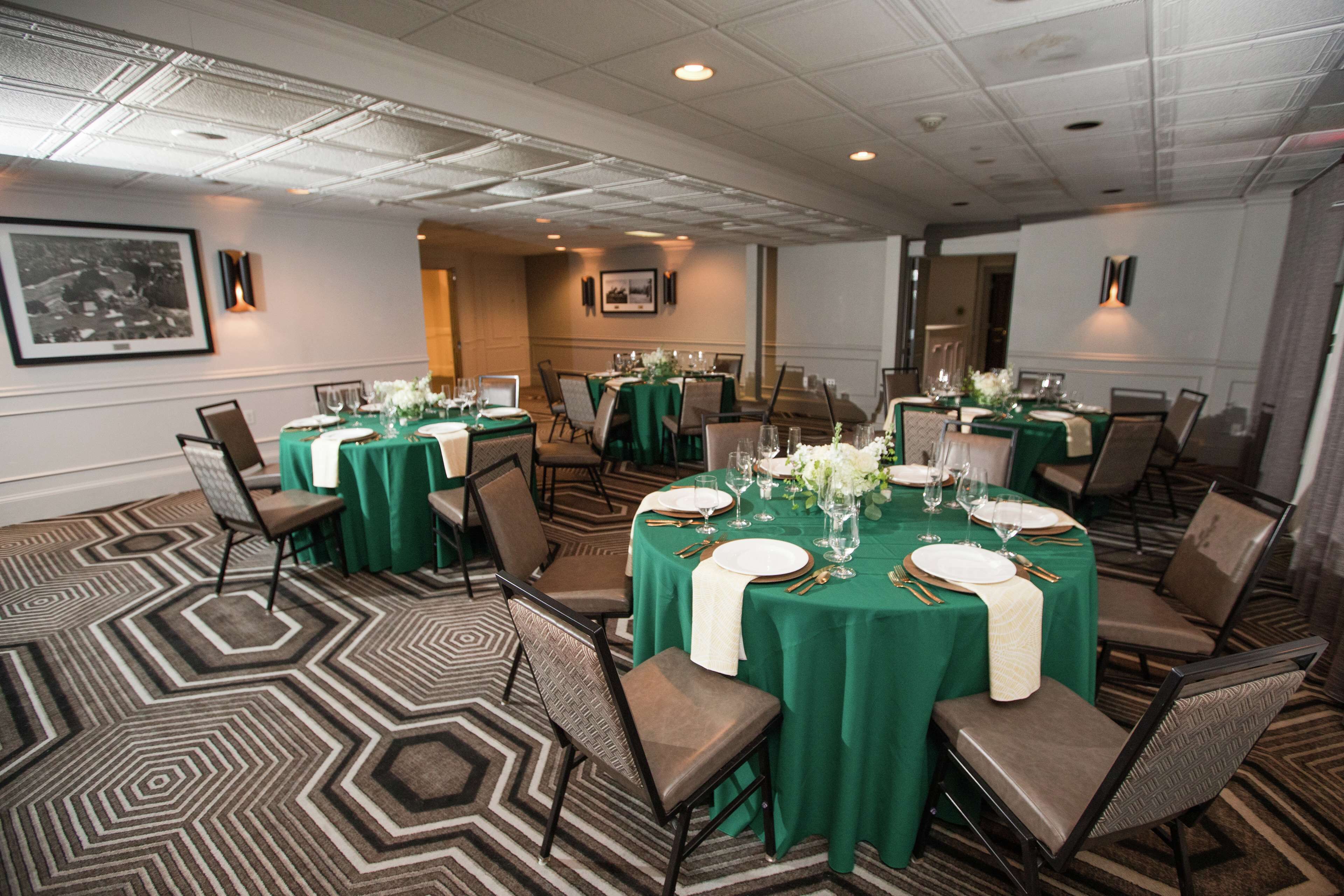 Un salón de conferencias con mesas dispuestas para un evento formal, con manteles verdes, platos blancos y cubiertos dorados.
