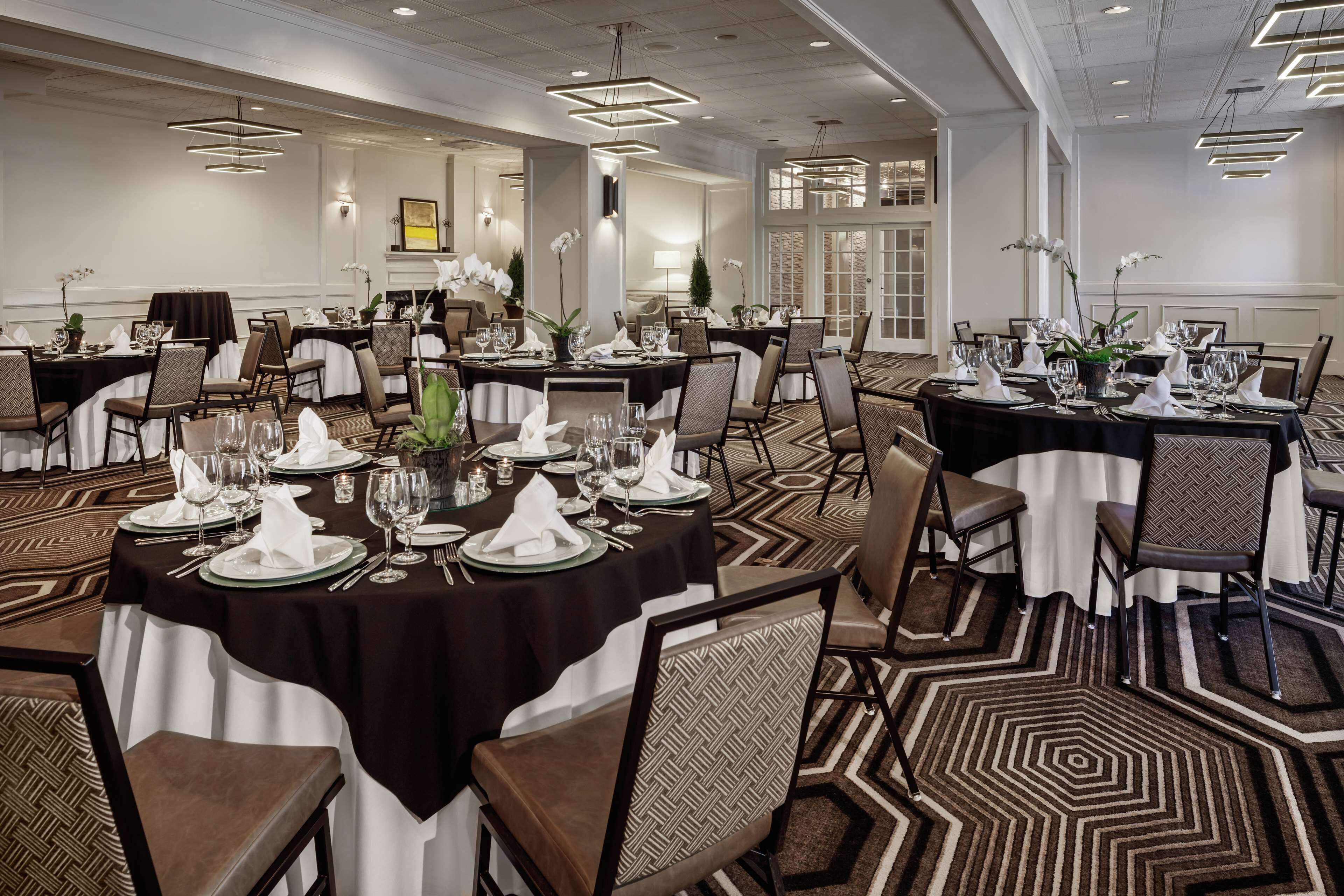 A large banquet hall with multiple round tables set for dining, featuring white tablecloths and neatly arranged place settings.