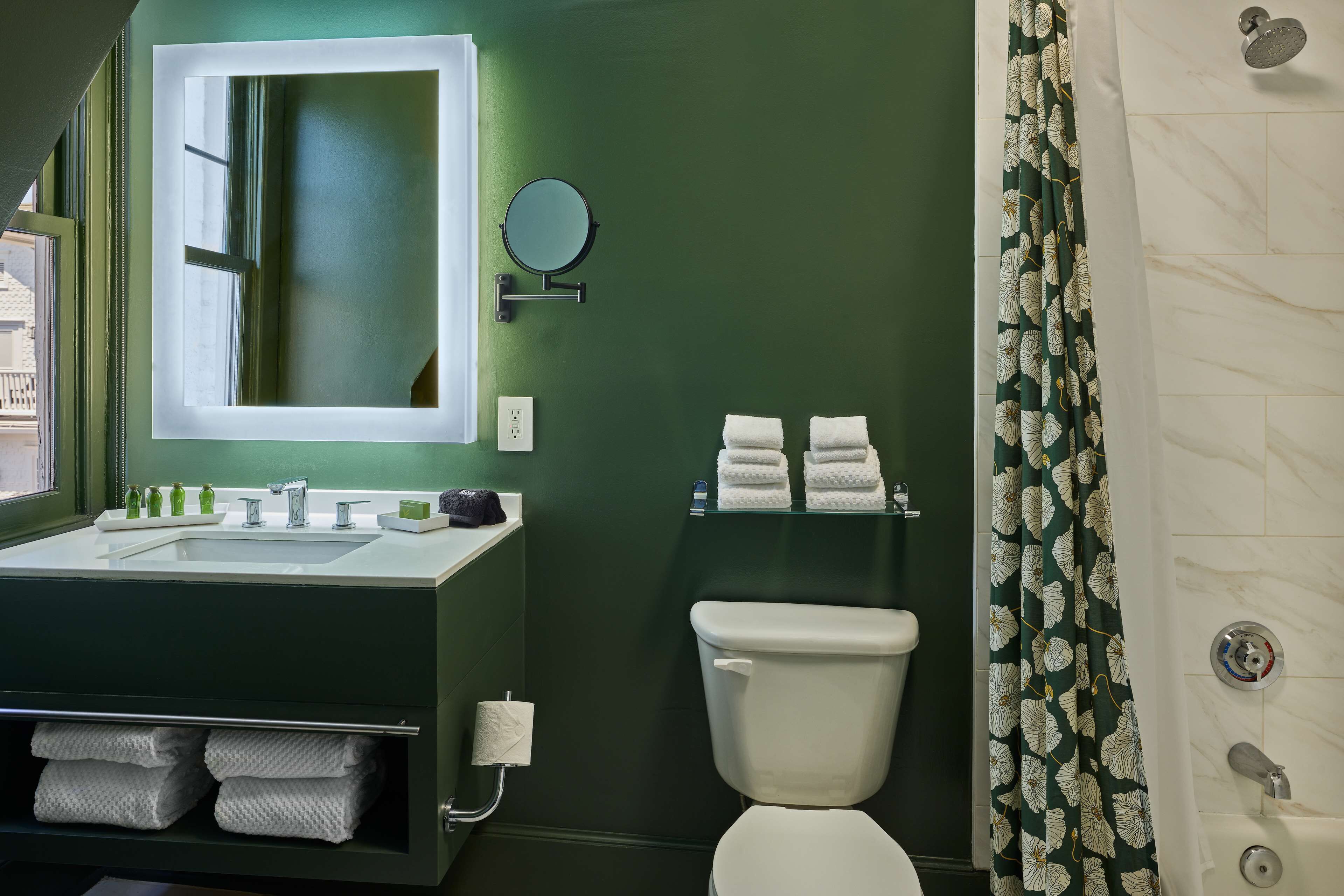 Un baño con pared verde, lavabo blanco y inodoro. Hay un espejo sobre el lavabo, una cortina de ducha y un perchero con toallas.