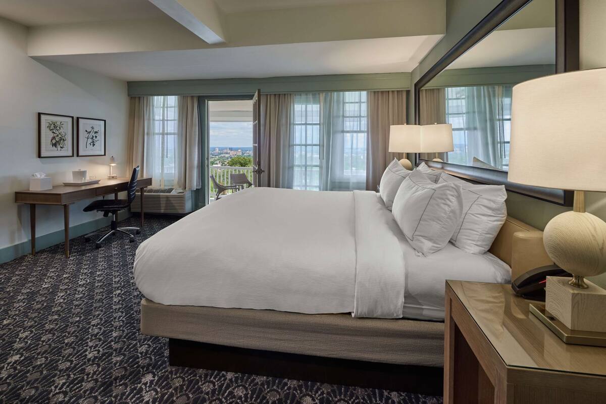 A hotel room with a large bed, a desk, a chair, a lamp, and a view of the city through the window.