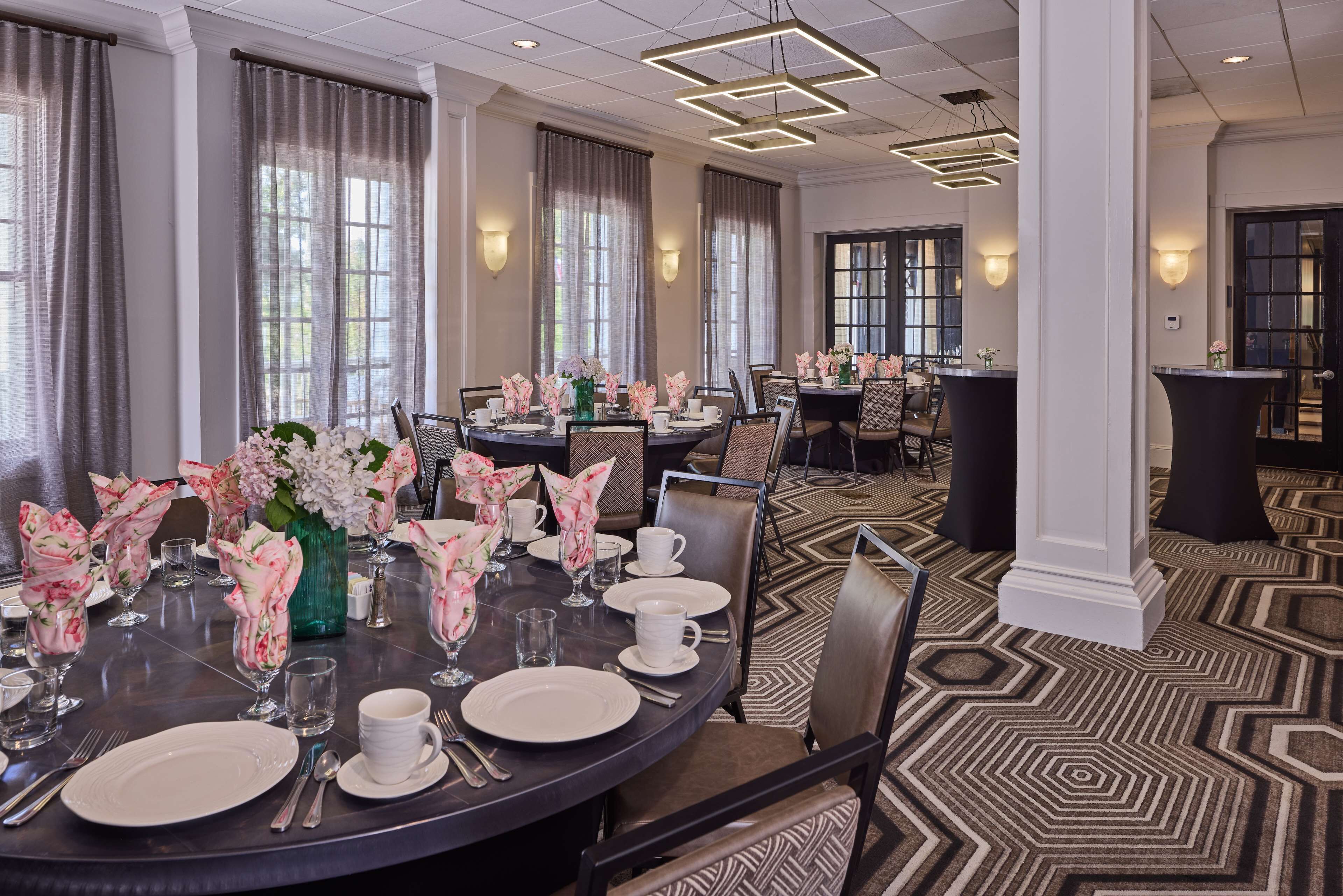 Un comedor de hotel con mesas redondas dispuestas para el servicio, ventanas amplias con cortinas y una alfombra estampada.