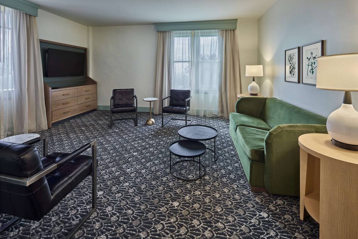 A hotel room with a green sofa, two armchairs, a small table, a TV on a wooden cabinet, and a patterned carpet.
