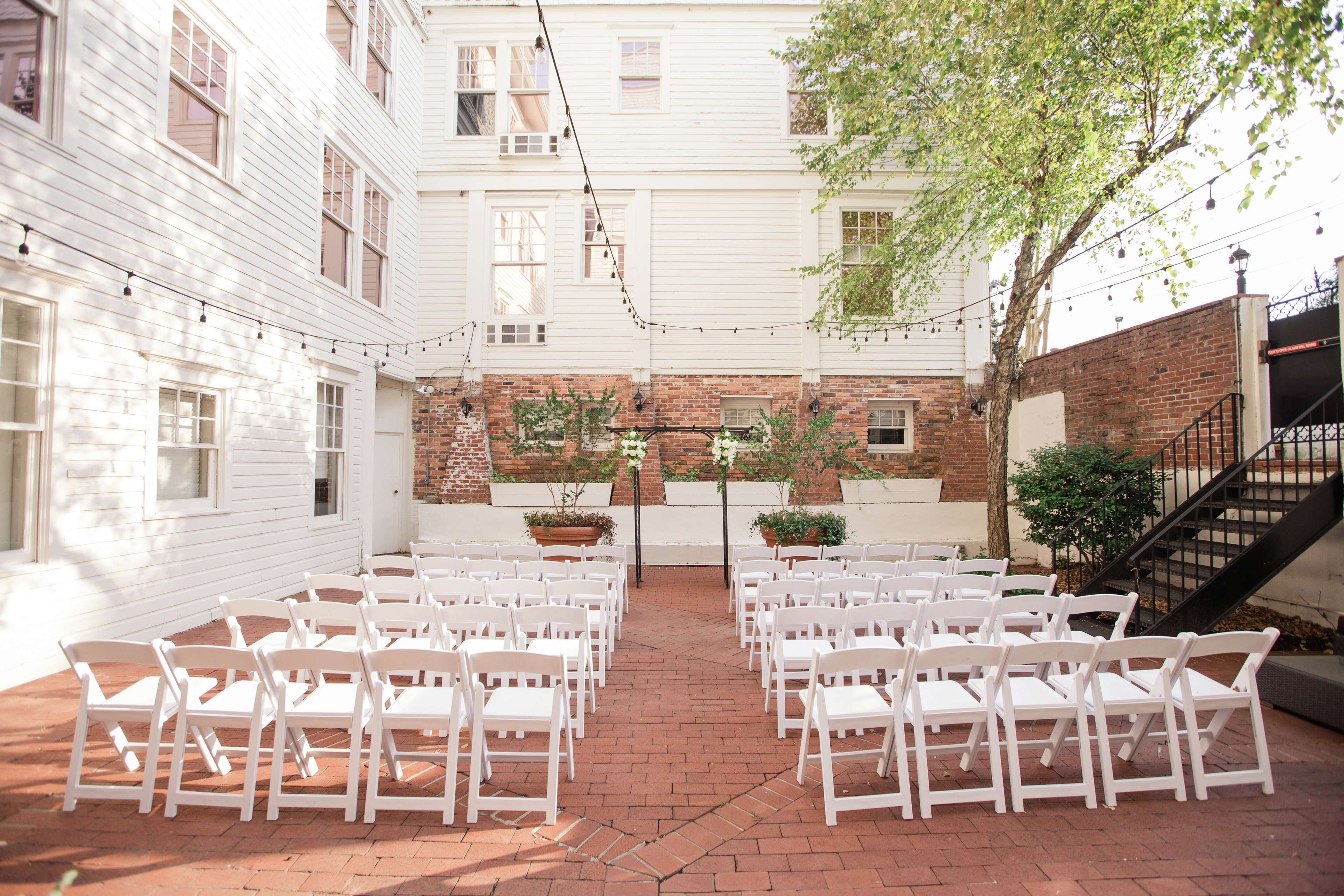 A courtyard with white chairs arranged for an event, surrounded by white buildings and a brick wall.