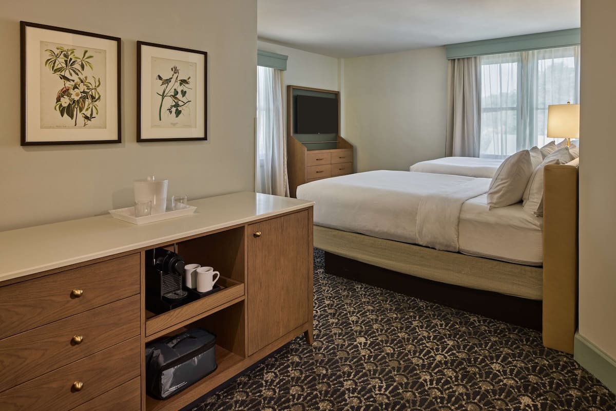 A hotel room with two beds, a TV, a desk with a lamp, and framed botanical prints on the wall.