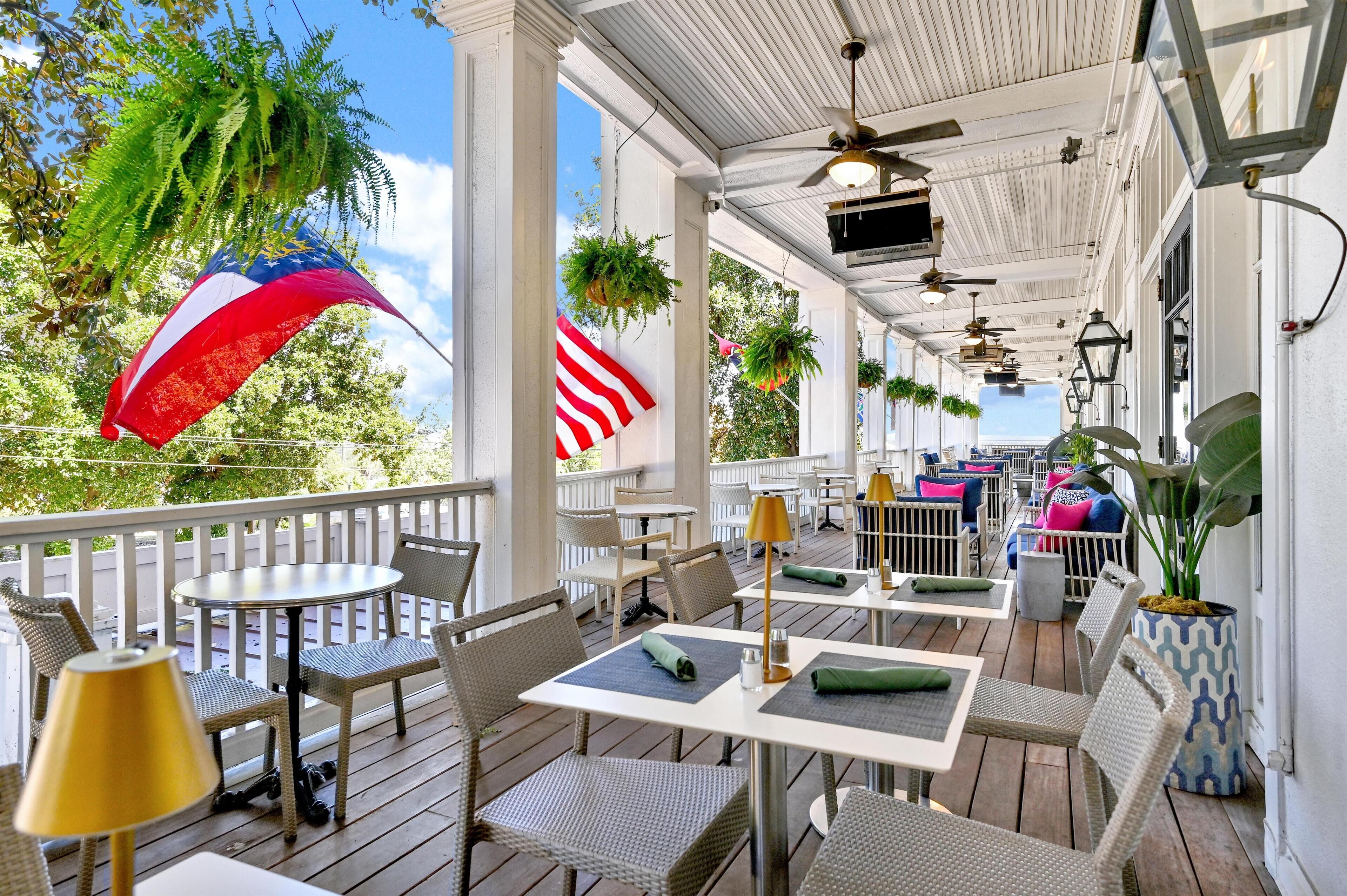A covered outdoor patio with tables and chairs, American flags, hanging plants, and a ceiling fan.