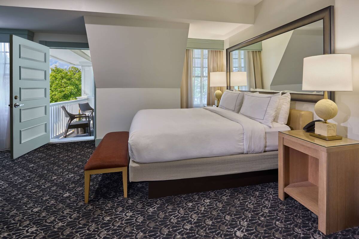 A hotel room with a bed, a bench, a nightstand with a lamp, a large mirror, and a view of the outdoors through an open door.