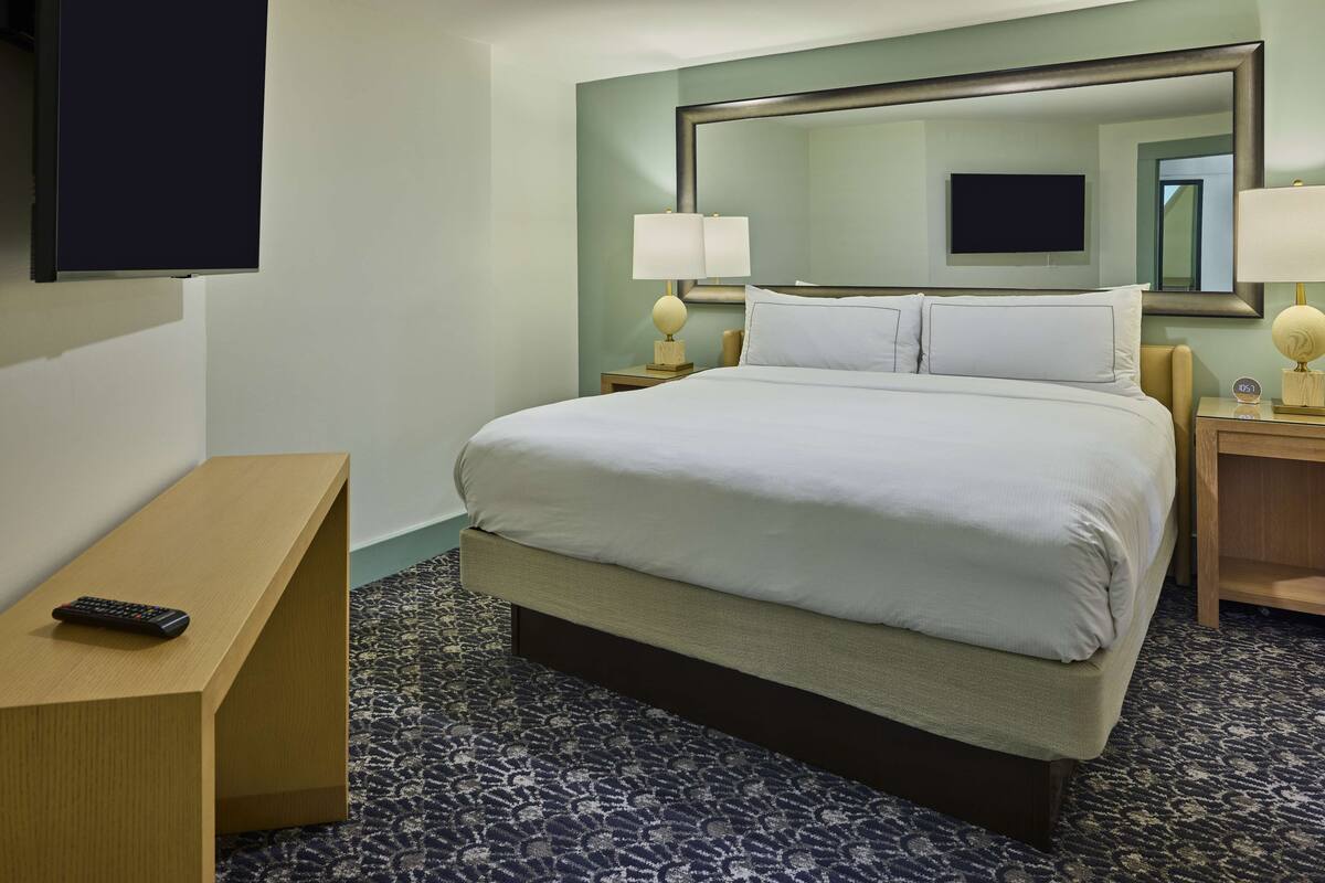 A hotel room with a large bed, a bedside table with a remote control, a nightstand with a lamp, and a large mirror above the bed.